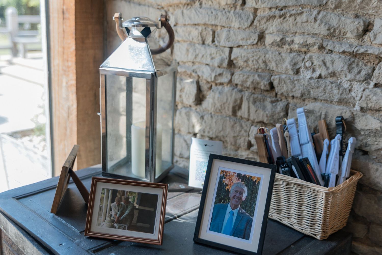 Silver lantern and framed photos displayed on dark table with wicker basket of pens against rustic stone wall.