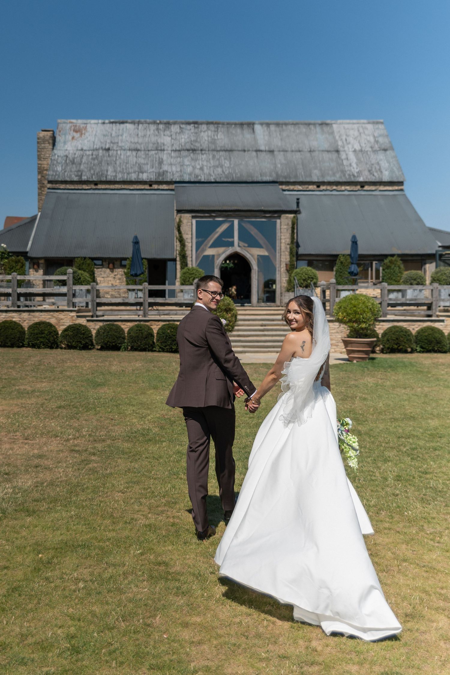 Bride and groom holding hands walking toward rustic wedding venue on sunny day.