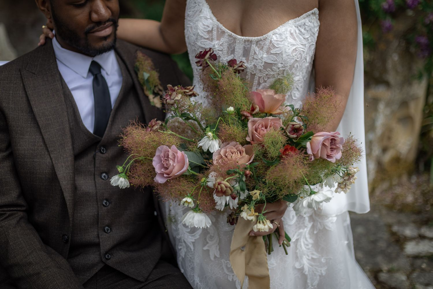 Couple on wedding day at Sudeley Castle, groom in brown suit, bride in lace gown holding a rustic pink floral bouquet.