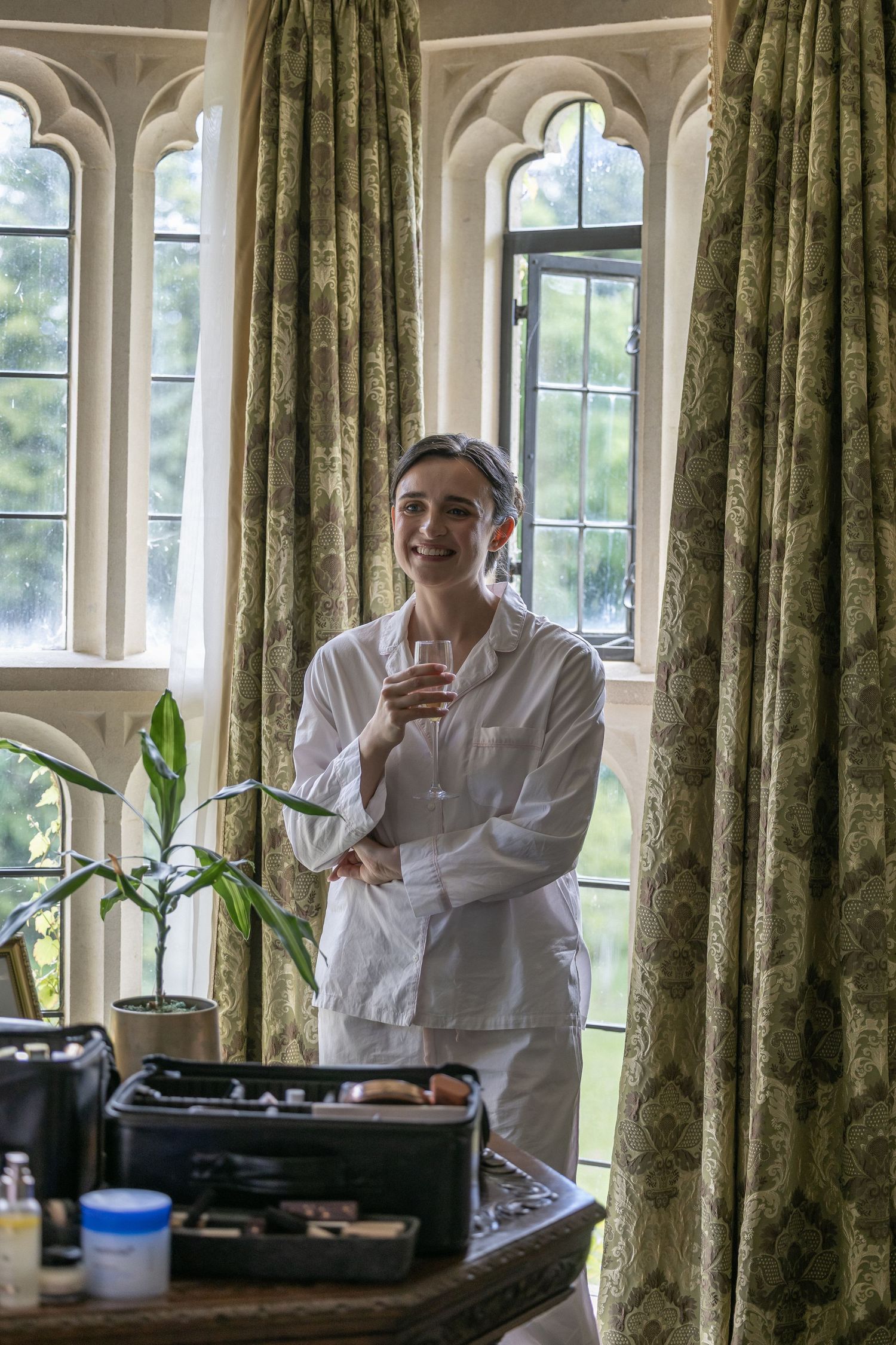 Smiling woman in white pajamas holding a drink by arched windows with green curtains and makeup kit.