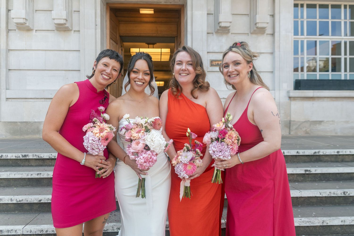 Bride in white gown poses with three bridesmaids in pink and red dresses holding floral bouquets on stone steps of Hackney Town Hall