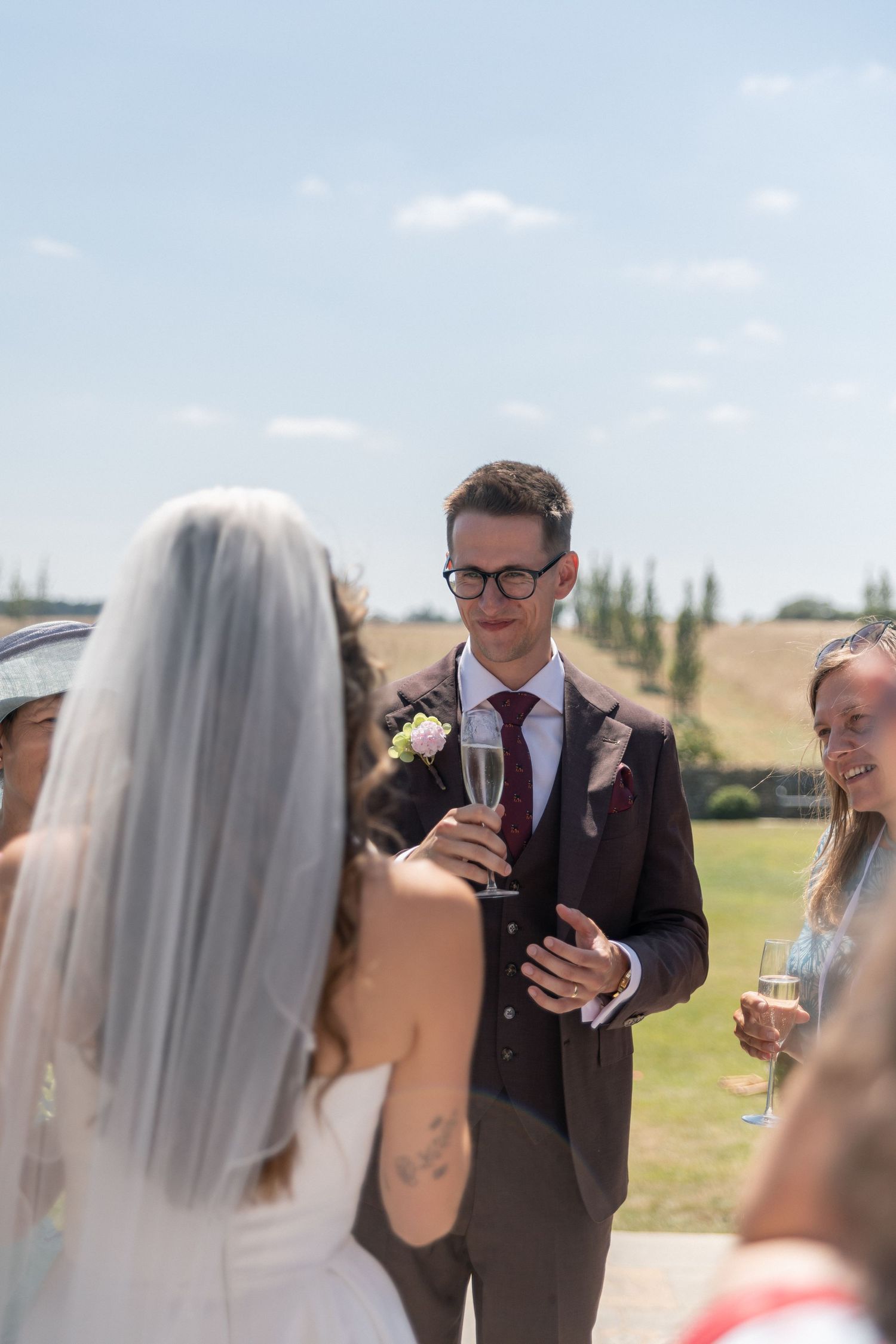 Groom in brown suit holding champagne glass toasts with bride in white dress and veil at outdoor wedding.
