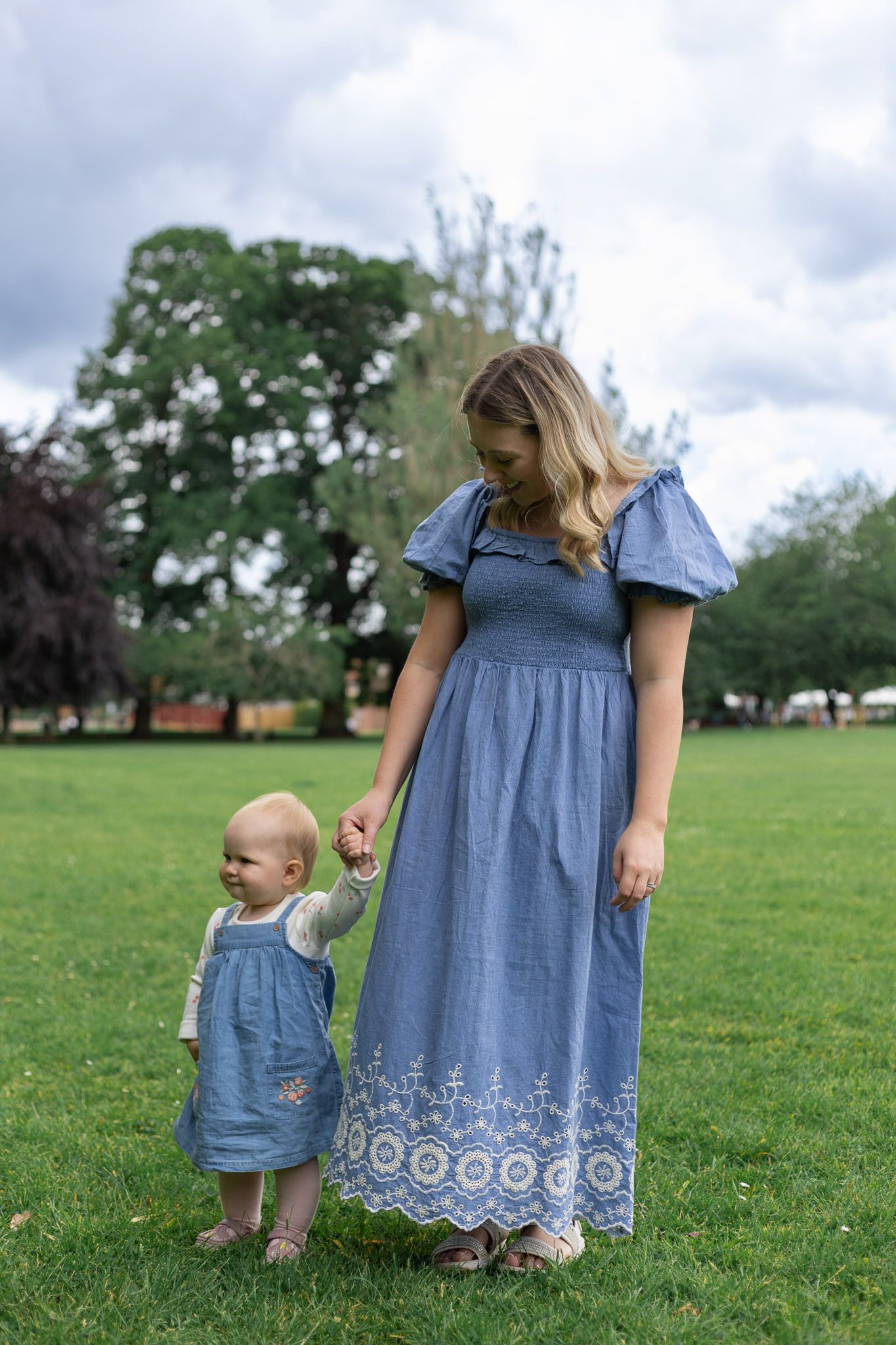Mother and child in matching blue dresses stand together on grassy field under cloudy sky.