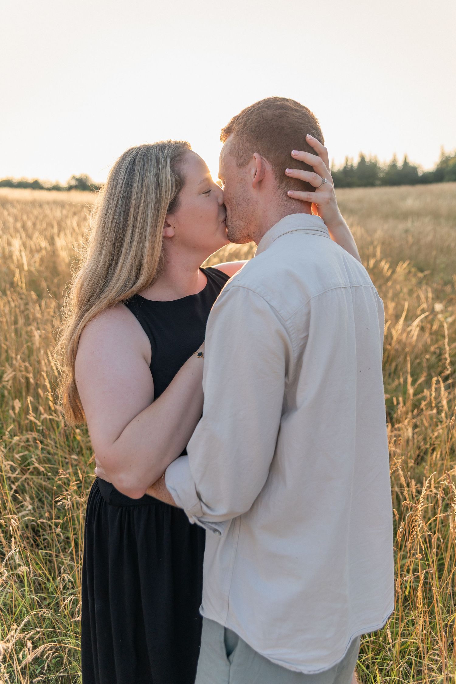 Couple sharing a romantic kiss in a golden wheat field at sunset, woman in black dress embracing man in white shirt.