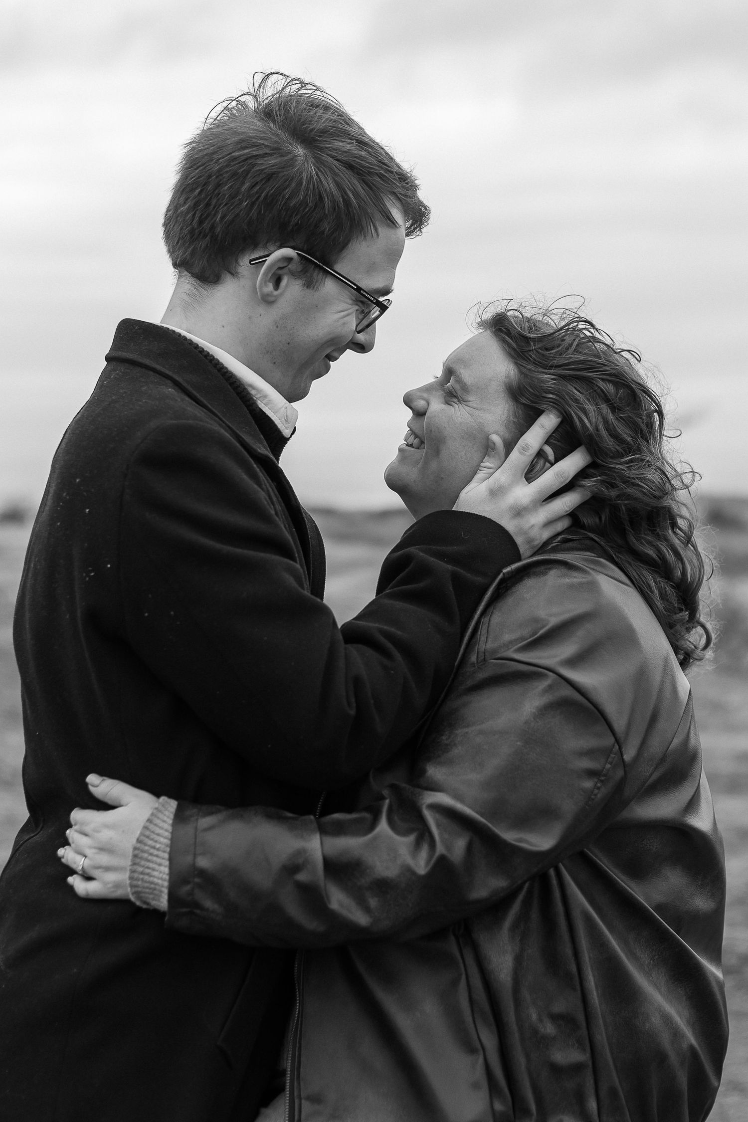 Black and white photo of a couple embracing and smiling at each other outdoors on a cloudy day.