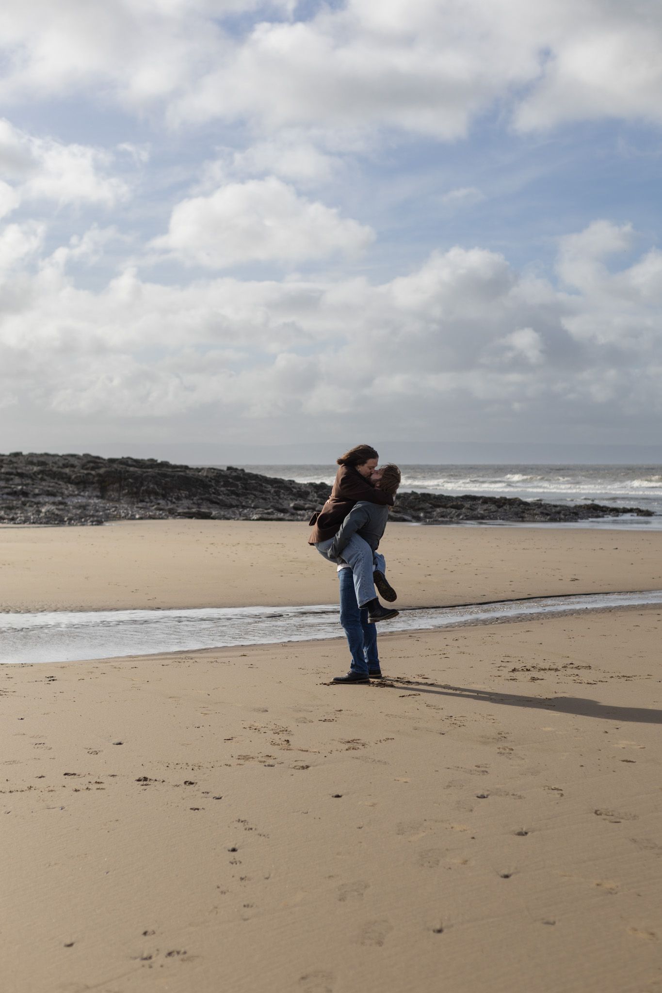 Two figures walking and playing on an empty beach with dramatic cloudy skies.