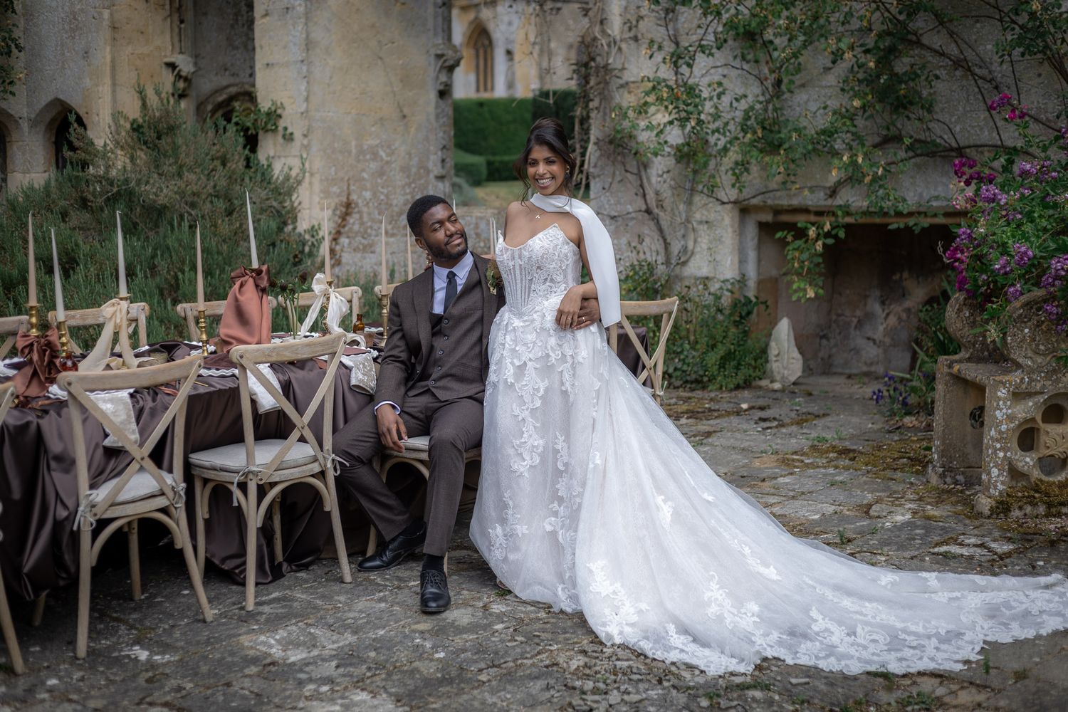 Elegant bride in lace gown and groom in gray suit pose at decorated table in rustic stone courtyard at Sudeley Castle