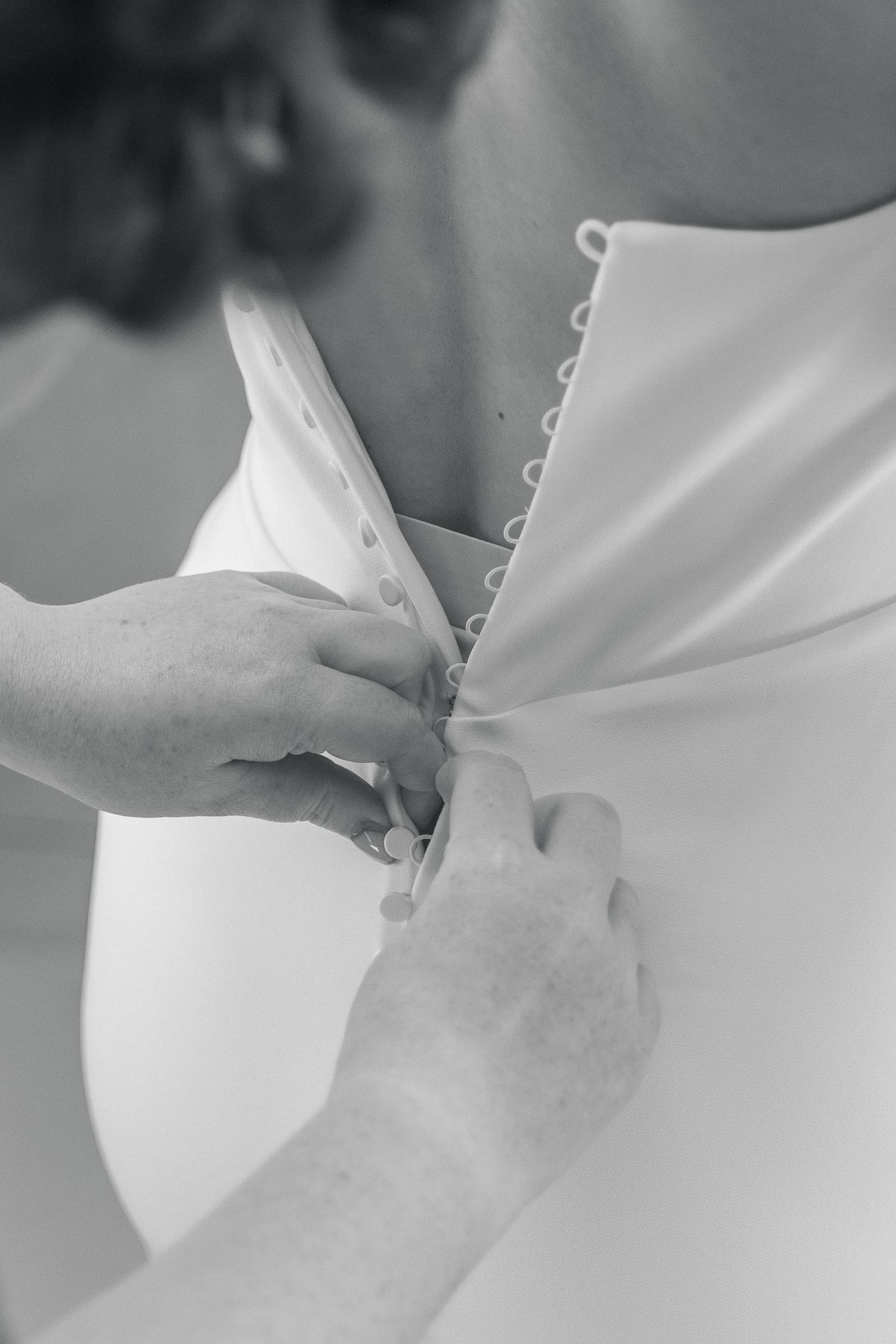 Black and white photo of hands fastening buttons on the back of a white wedding dress.