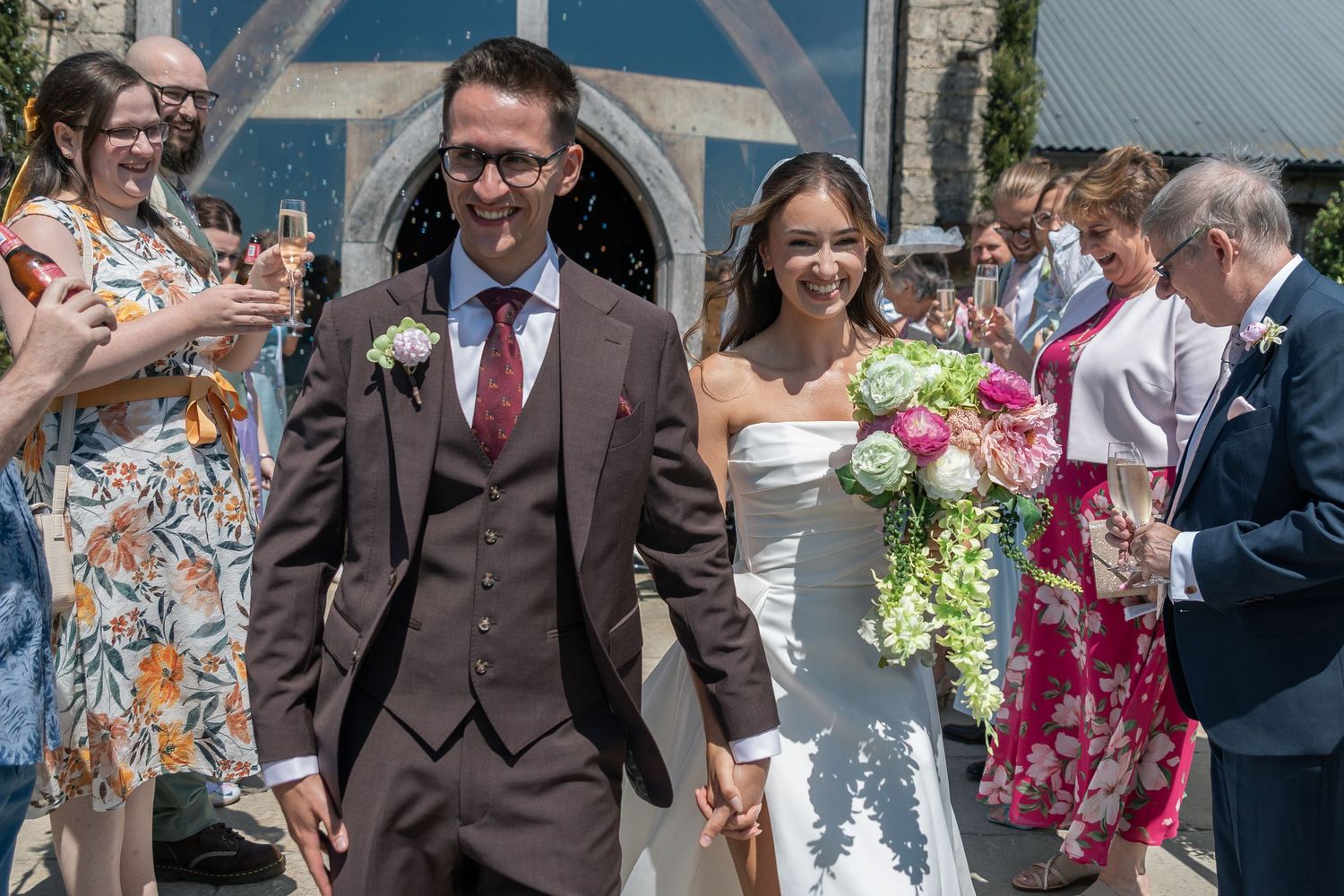 Smiling newlyweds walk hand-in-hand through confetti shower outside stone church, bride holding pink bouquet.