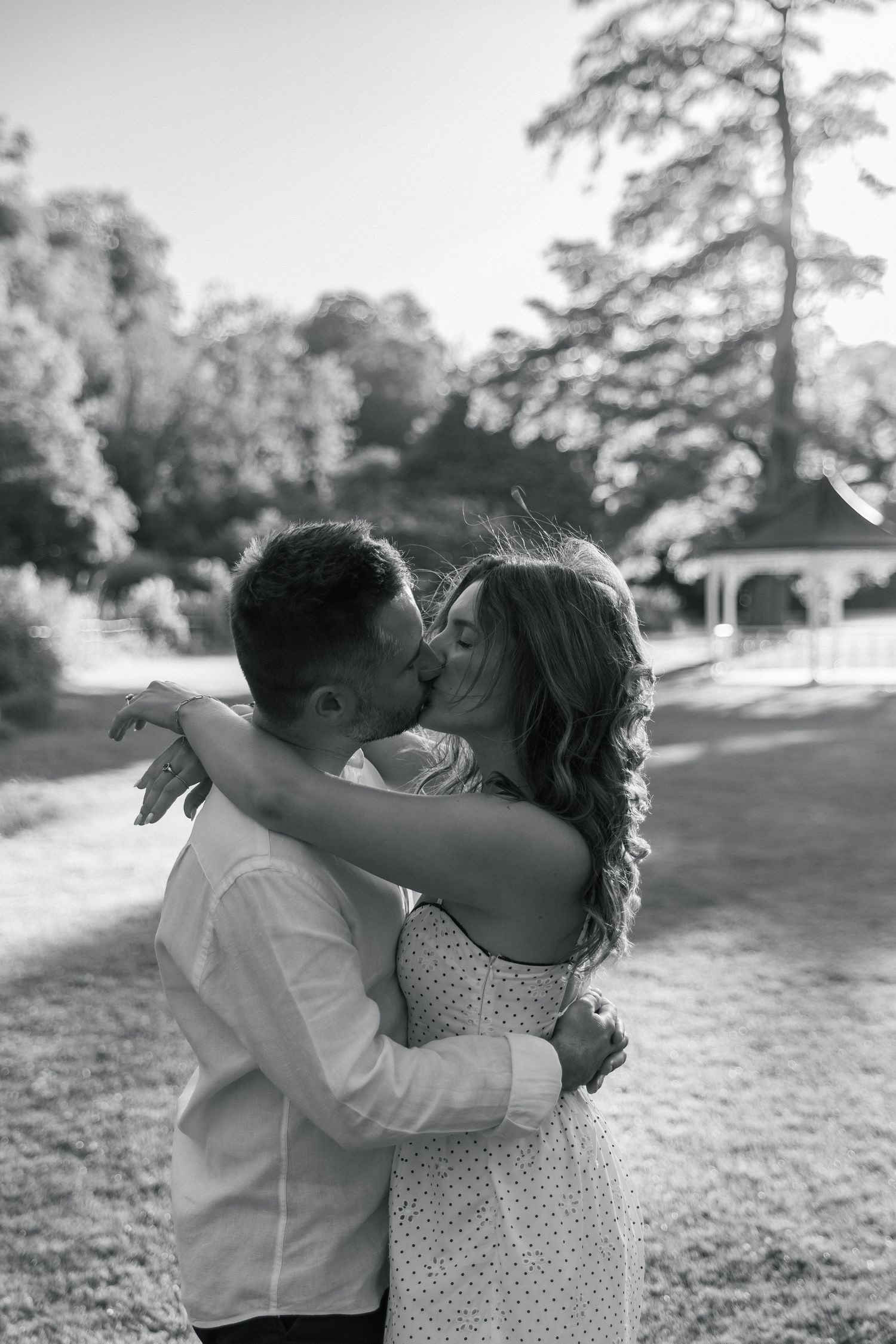 Black and white photo of a couple kissing and embracing outdoors in a park with a gazebo in the background.