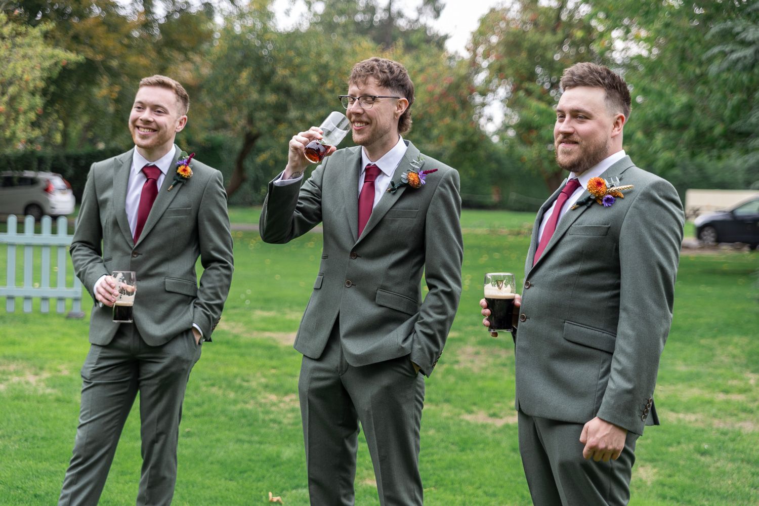 Groomsmen in matching gray suits and red ties laugh together on a lawn.