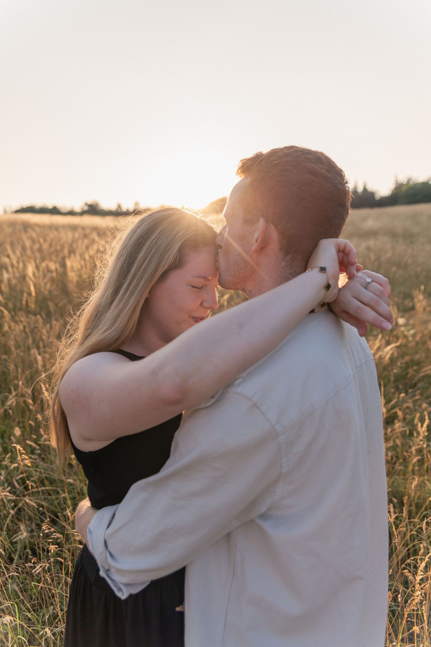 Couple embracing romantically in a golden wheat field at sunset, woman in black dress with arms around man's neck.