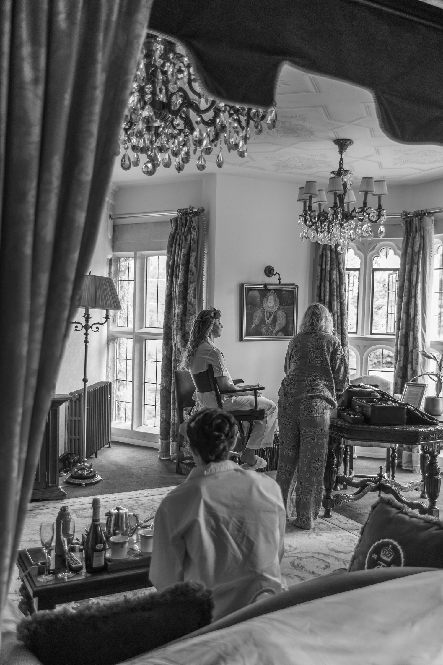 Black and white photo of women getting ready in an elegant room with chandeliers and four-poster bed.