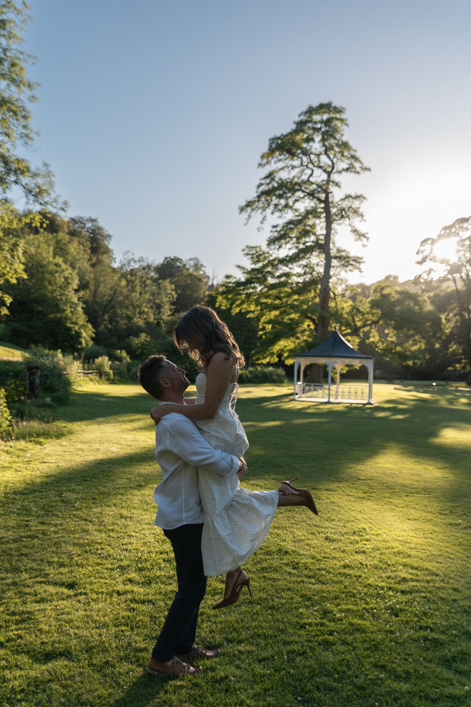 Man lifting woman in white dress on lush green lawn with gazebo and trees in golden sunset light.