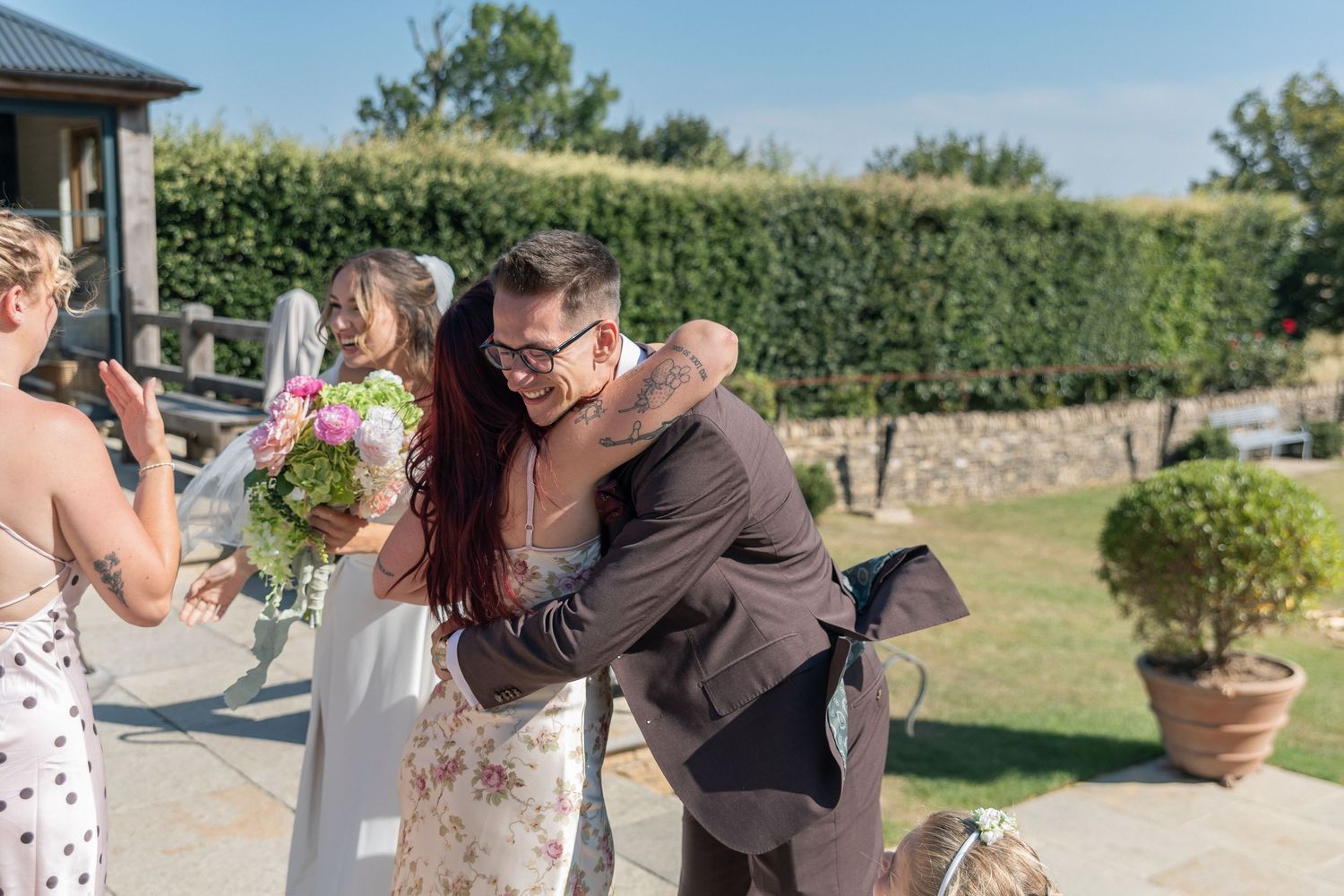 Bride and groom share a joyful hug outdoors at a sunny wedding venue surrounded by guests.