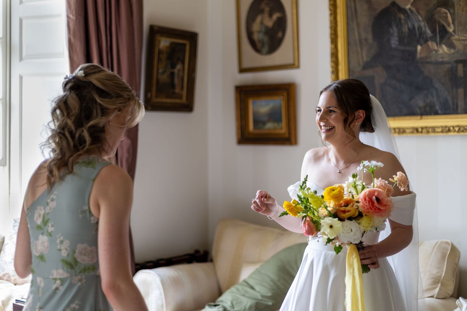 A bride and bridesmaids in soft pastel dresses prepare for a wedding ceremony in an elegant room with framed artwork.