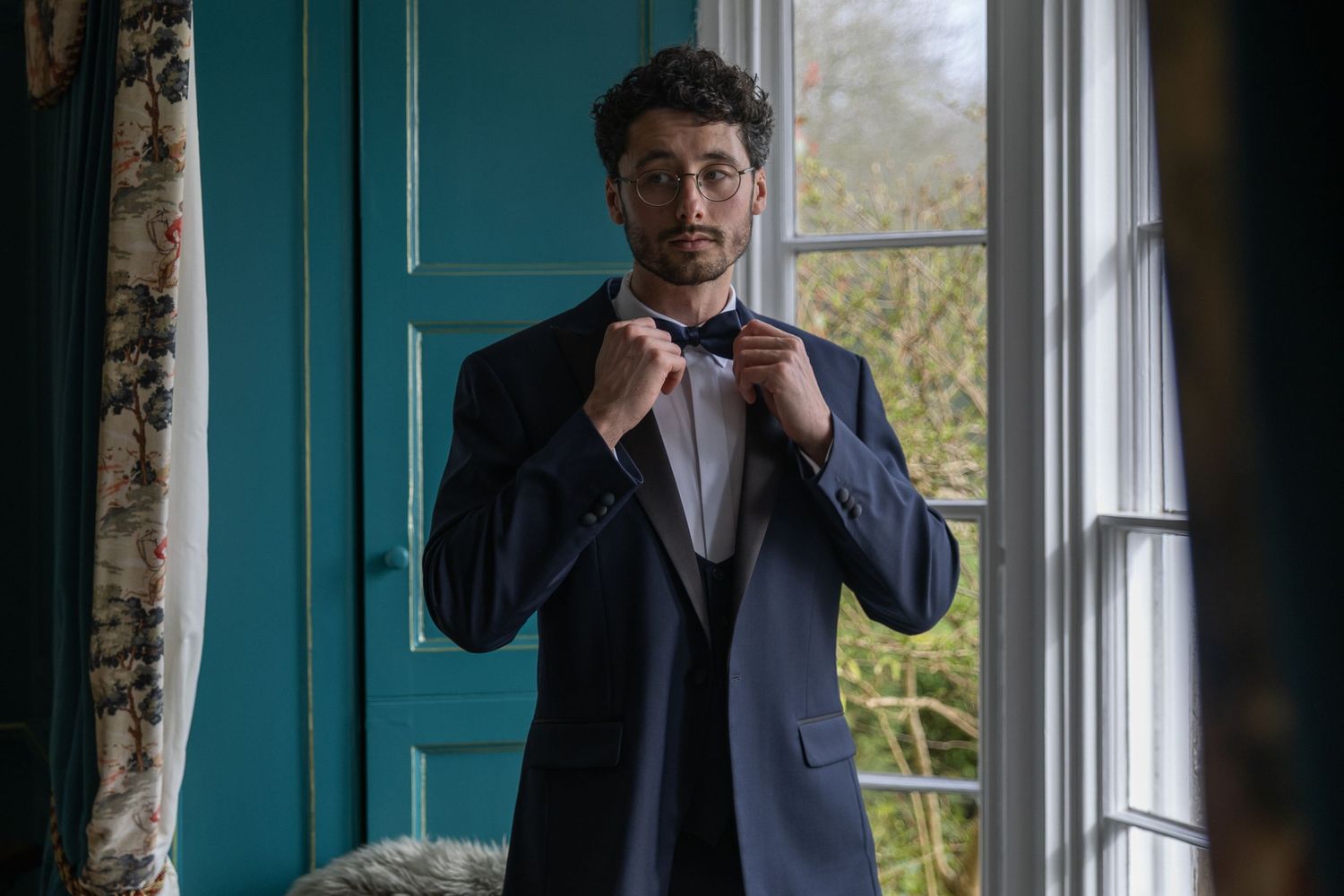 Stylish young man adjusting bow tie wearing navy blue suit jacket standing by teal window in elegant room.