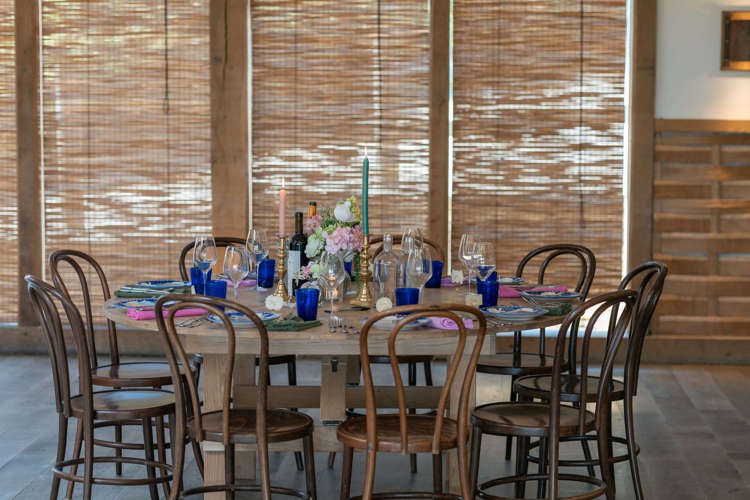 Round dining table with bentwood chairs, blue glassware, candles, and floral centerpiece set against bamboo blinds.