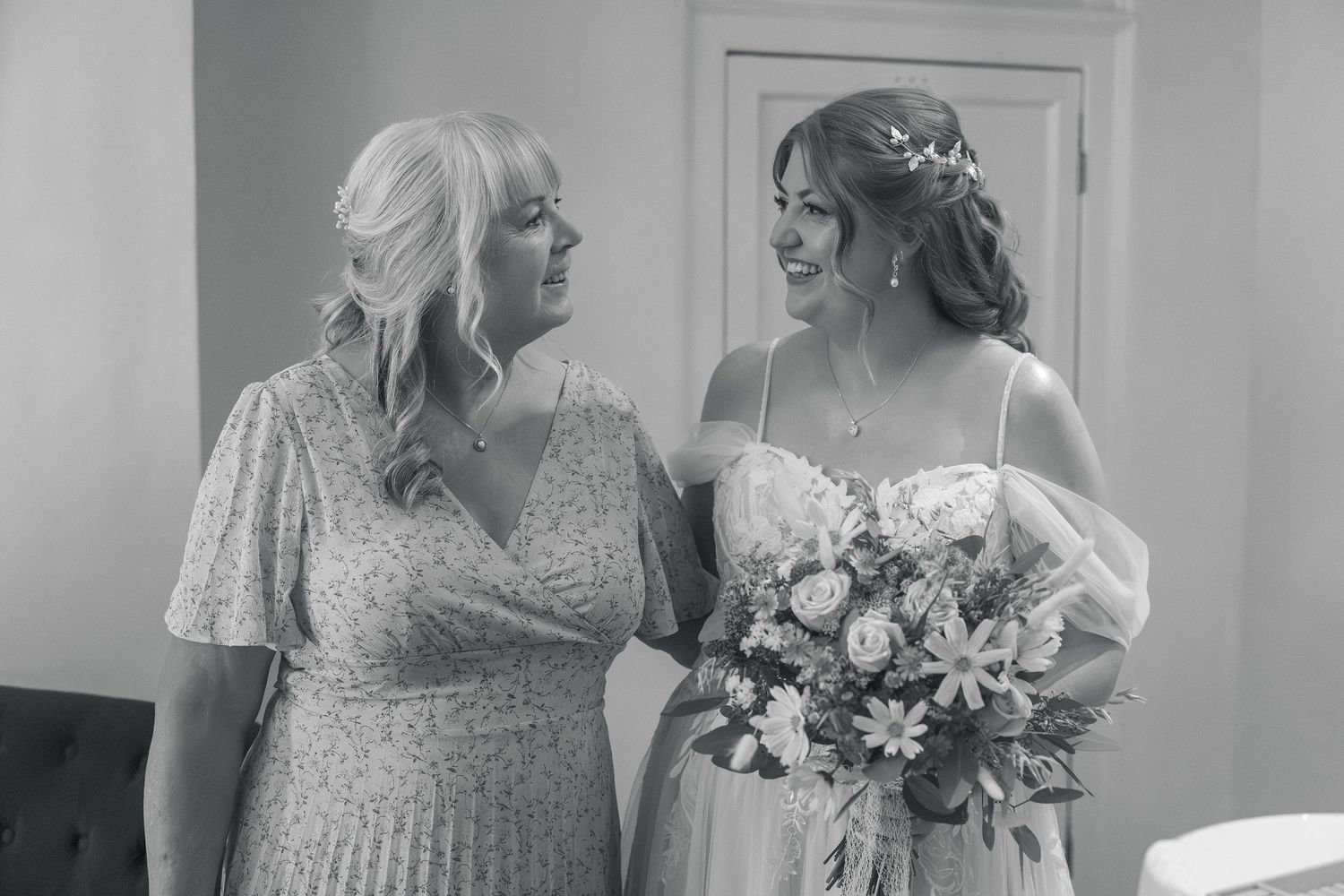Black and white photo of a bride holding a bouquet smiling with an older woman in a floral dress at a wedding.