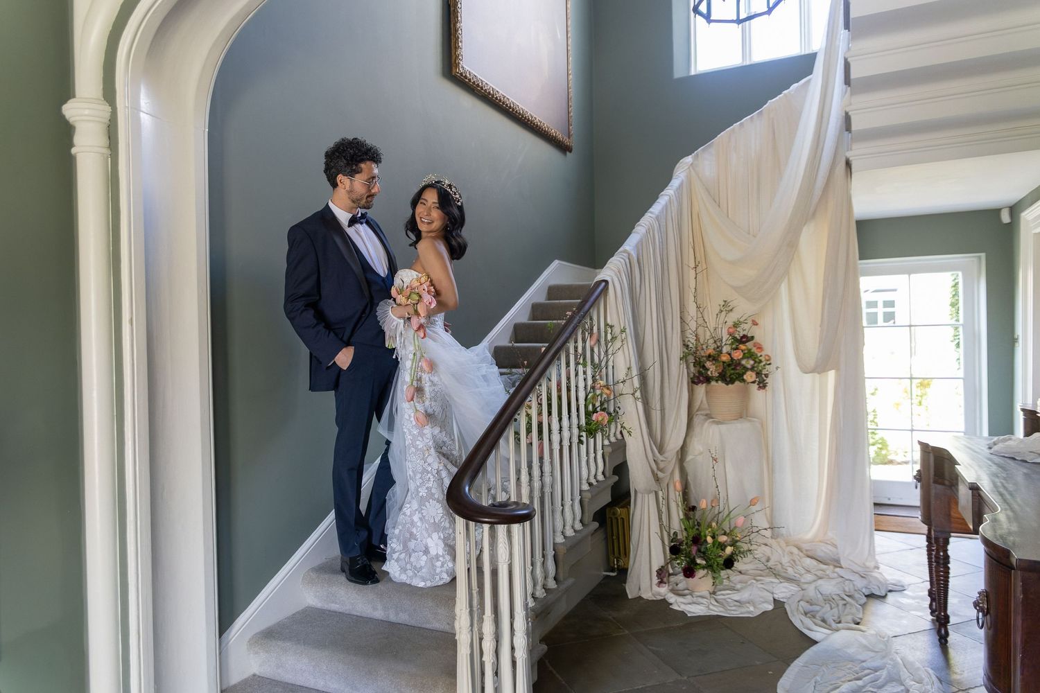 Bride and groom stand elegantly on a grand staircase in a decorated wedding venue with floral arrangements.