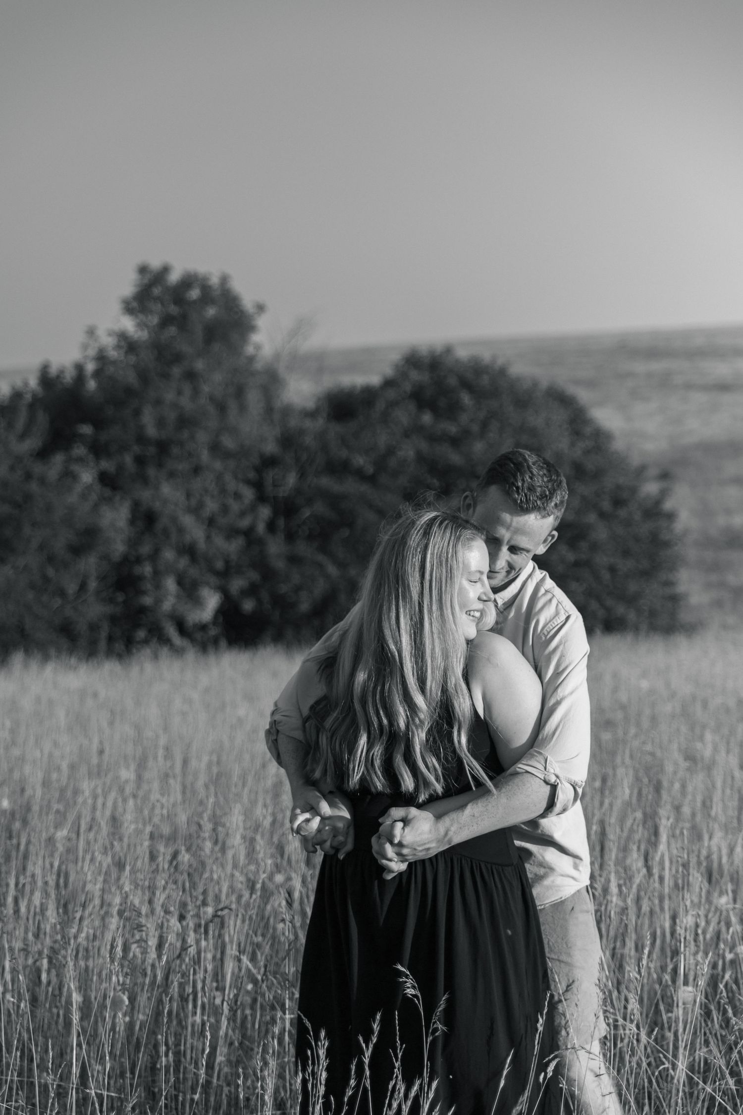 Black and white photo of a couple embracing in a grassy field with trees in the background.
