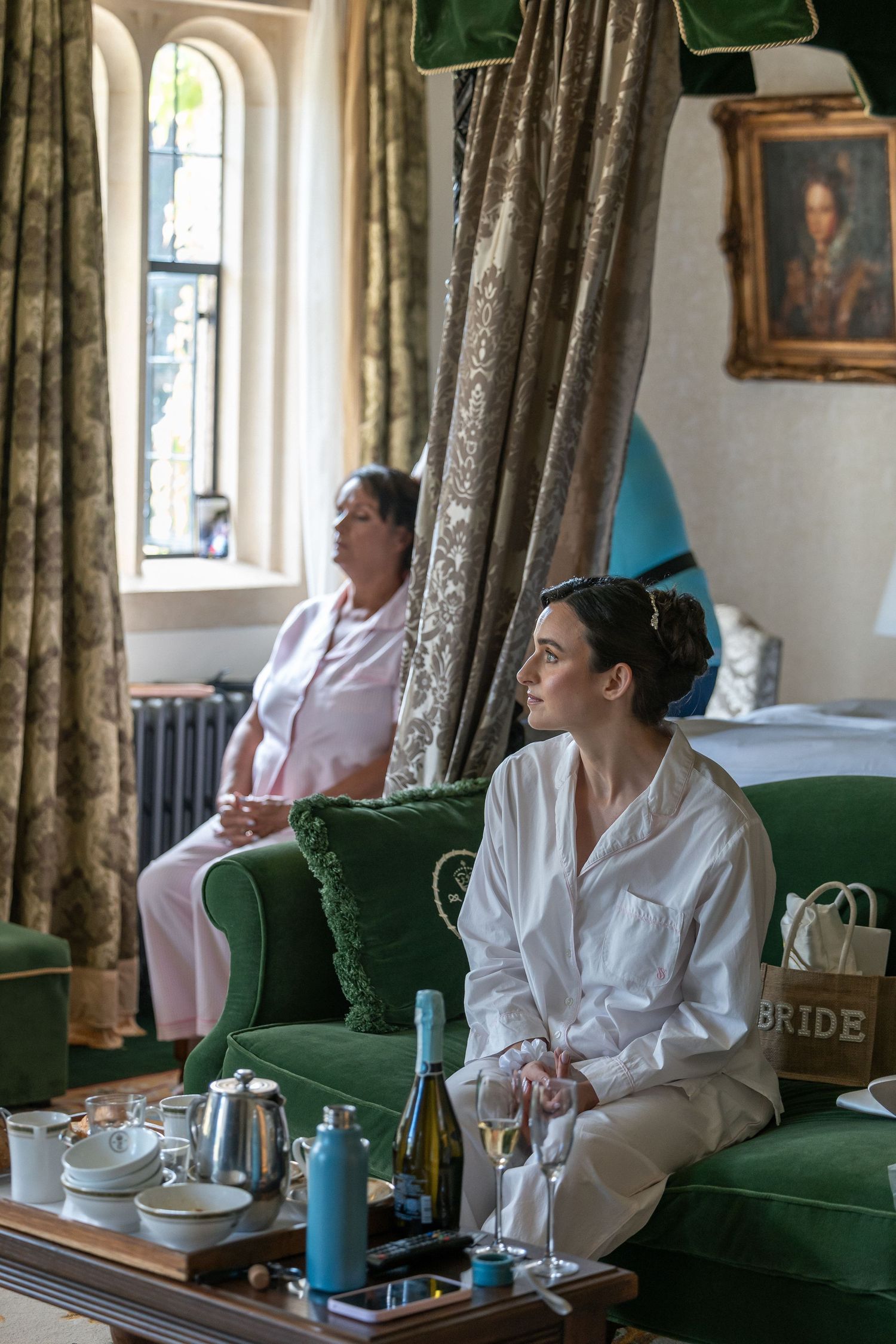 Bride in white robe sits on green velvet sofa with wine glass, BRIDE bag visible, elegant room with gold curtains.