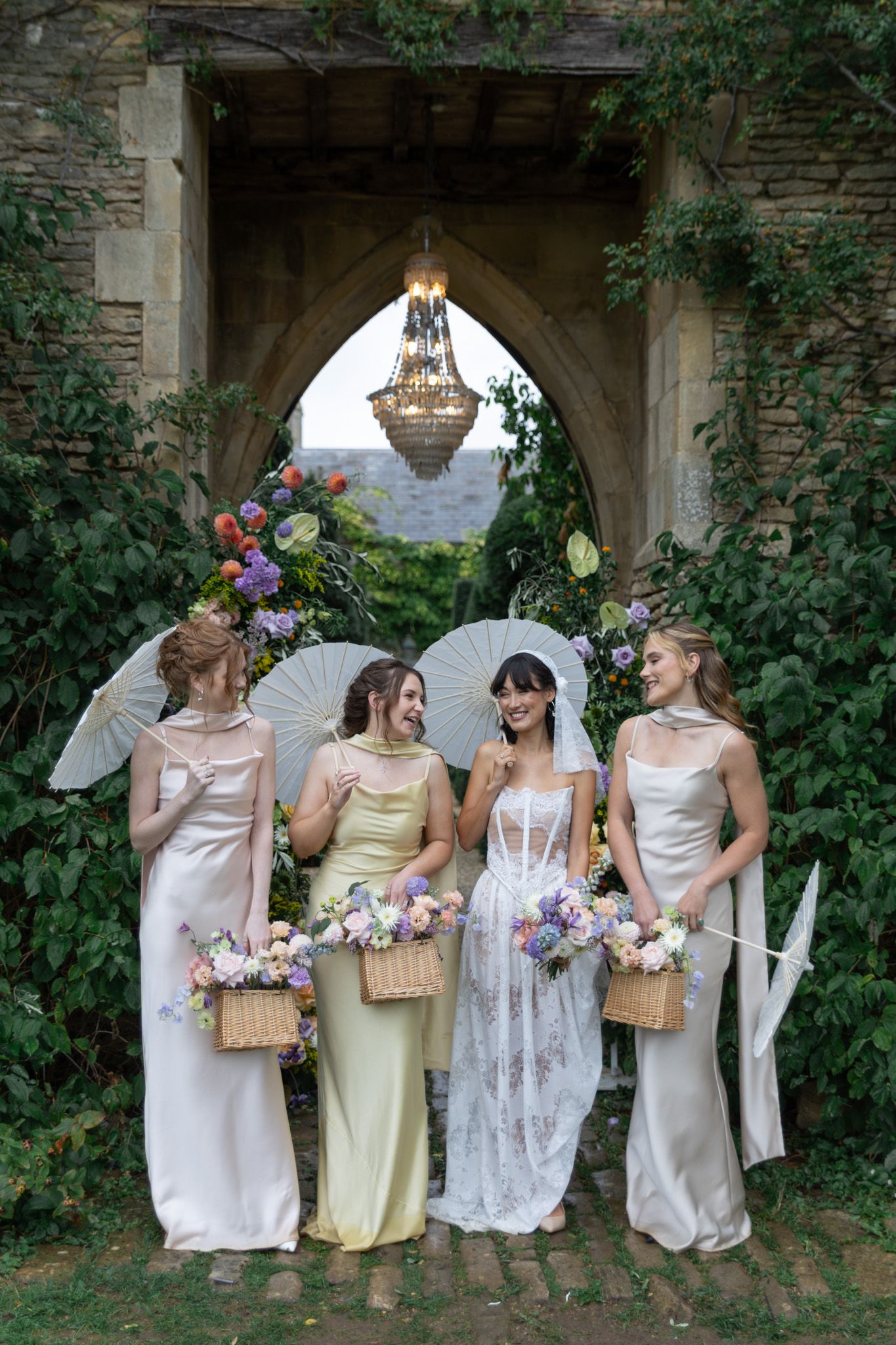Wedding party in elegant dresses poses against stone wall with greenery while holding wicker baskets and bouquets.