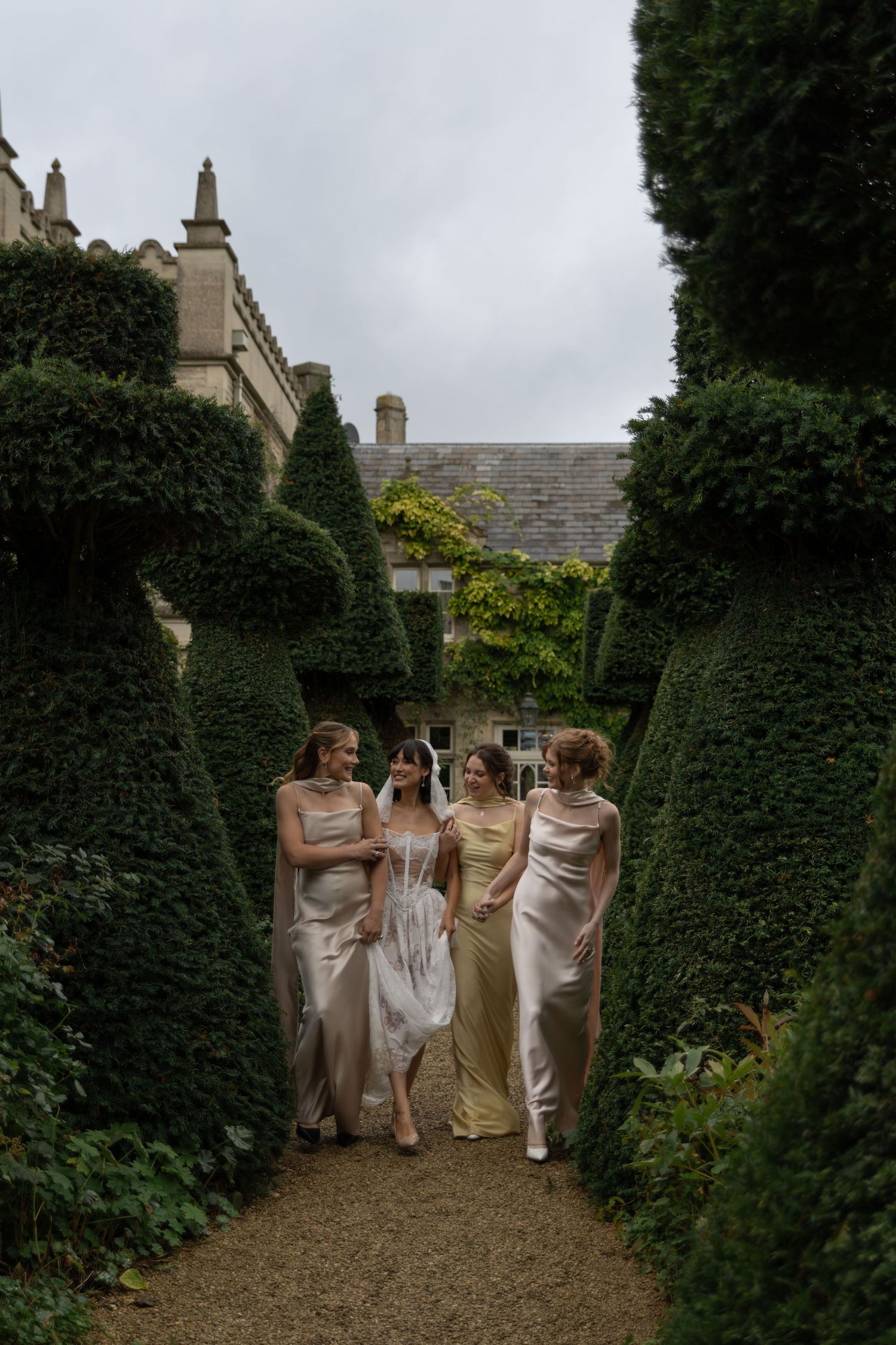 A bridal party in elegant champagne-colored dresses walks down a garden path lined with manicured hedges.