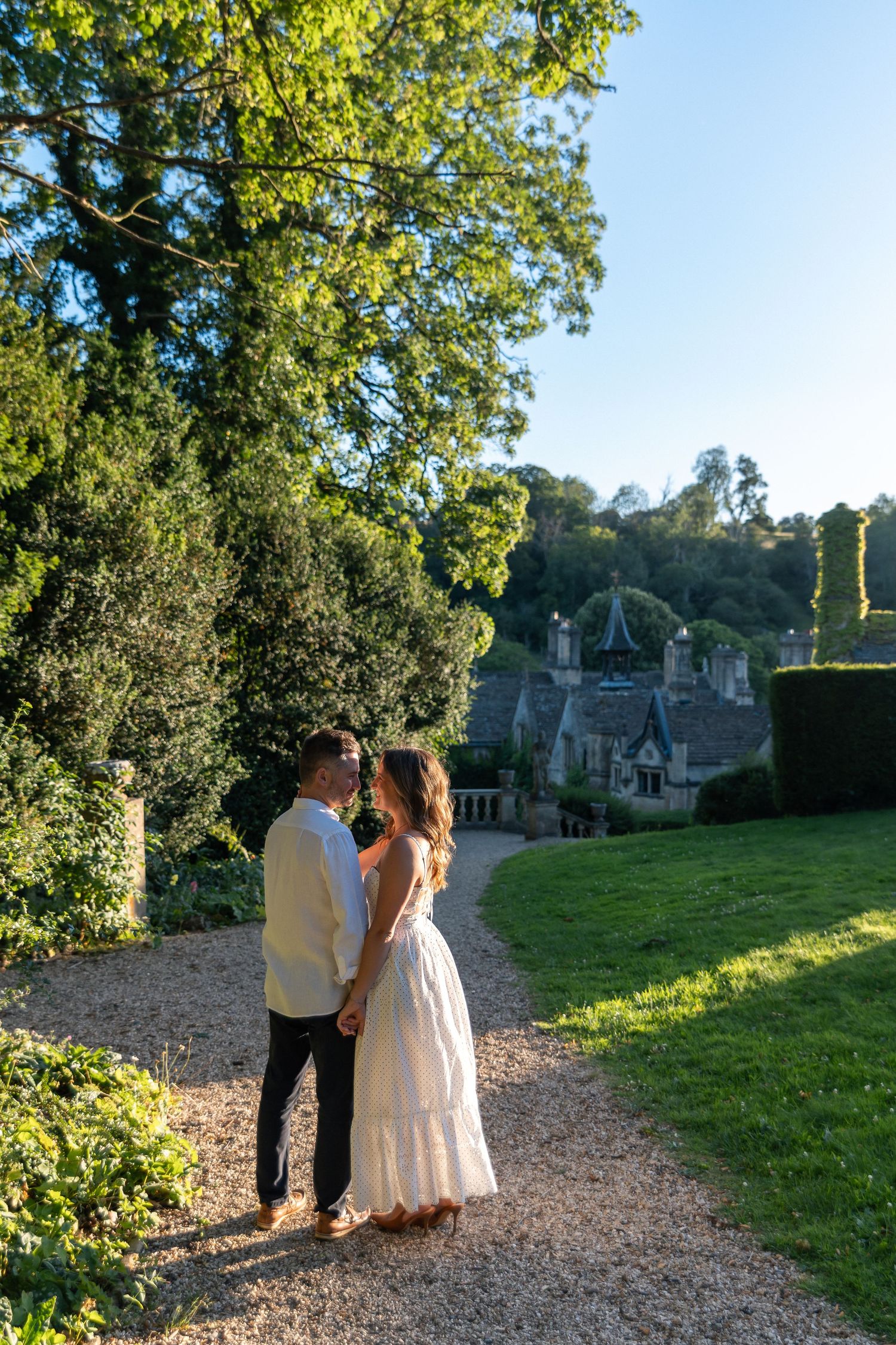 Couple holding hands on a gravel path in an English country garden with a stone manor house backdrop.