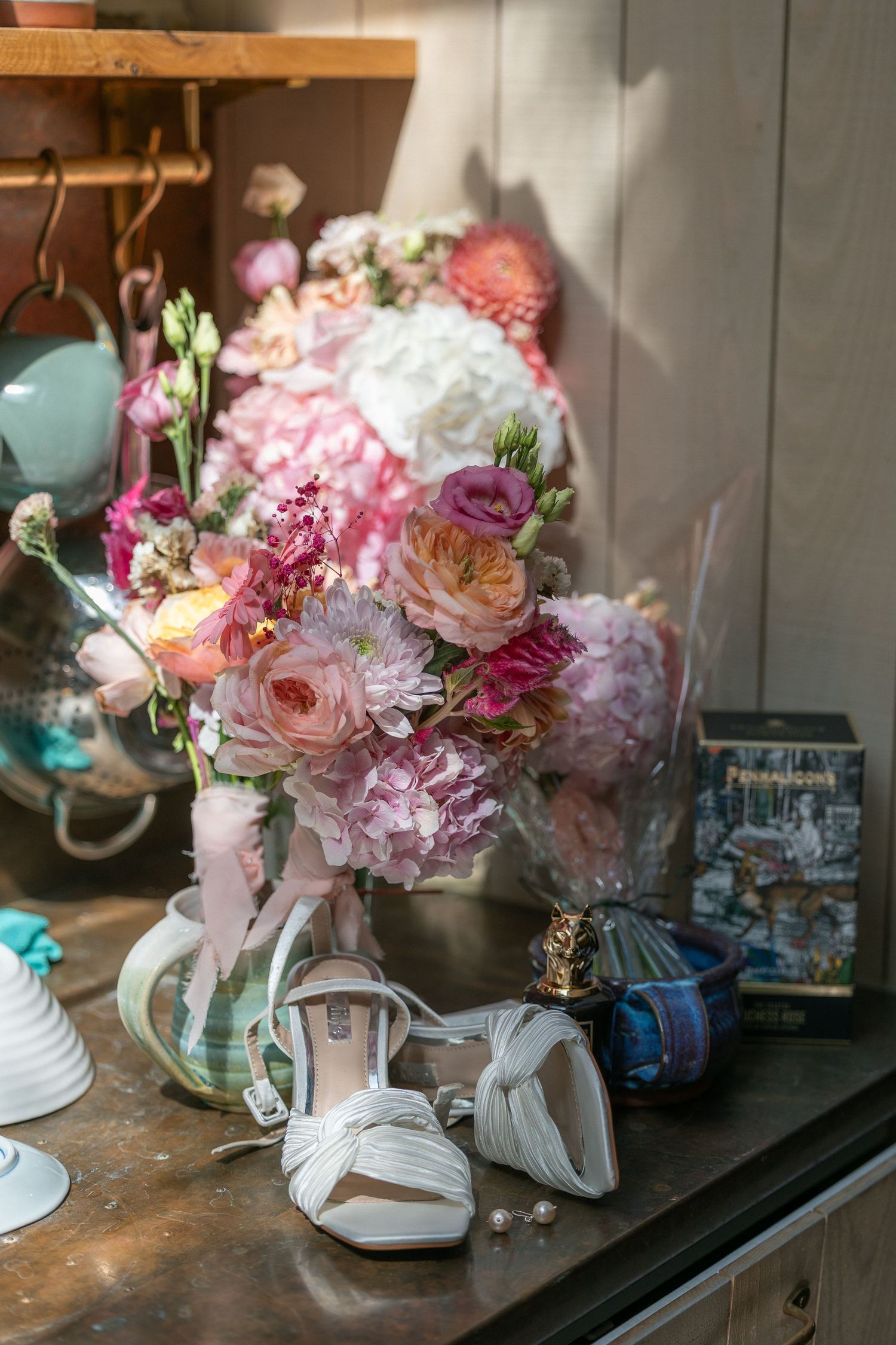 Bridal flat shoes beside a pink floral bouquet with peonies and roses, perfume bottle, and decorative items on a dark surface.