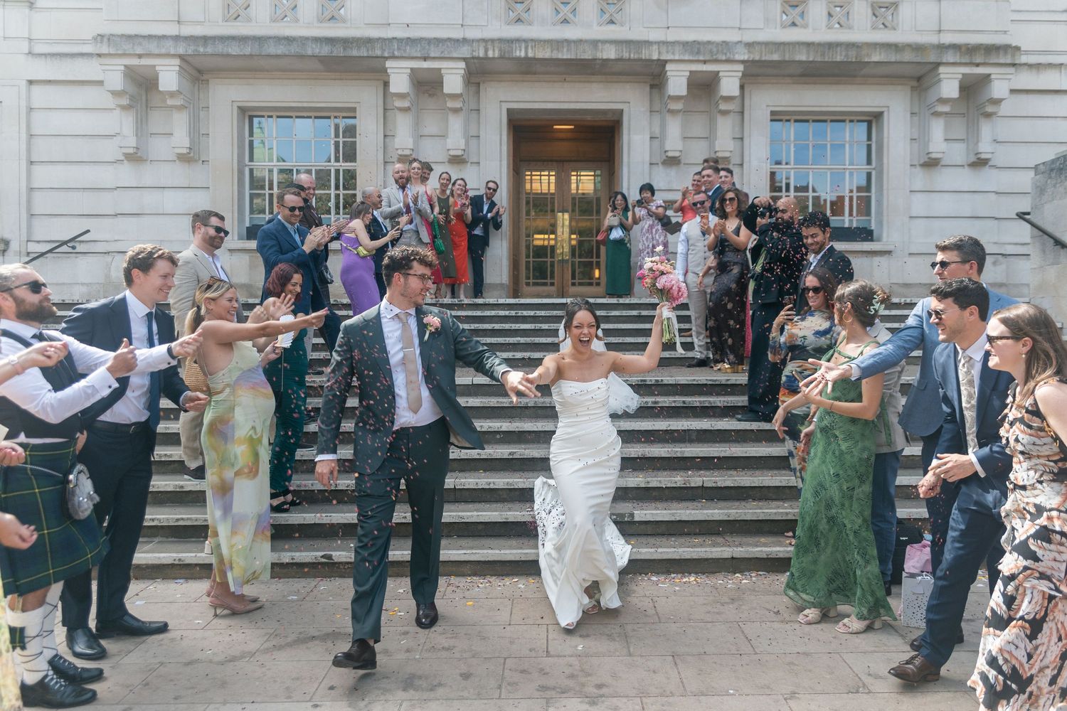 Bride and groom exit Hackney Town Hall on steps while guests celebrate with confetti in sunny outdoor ceremony.