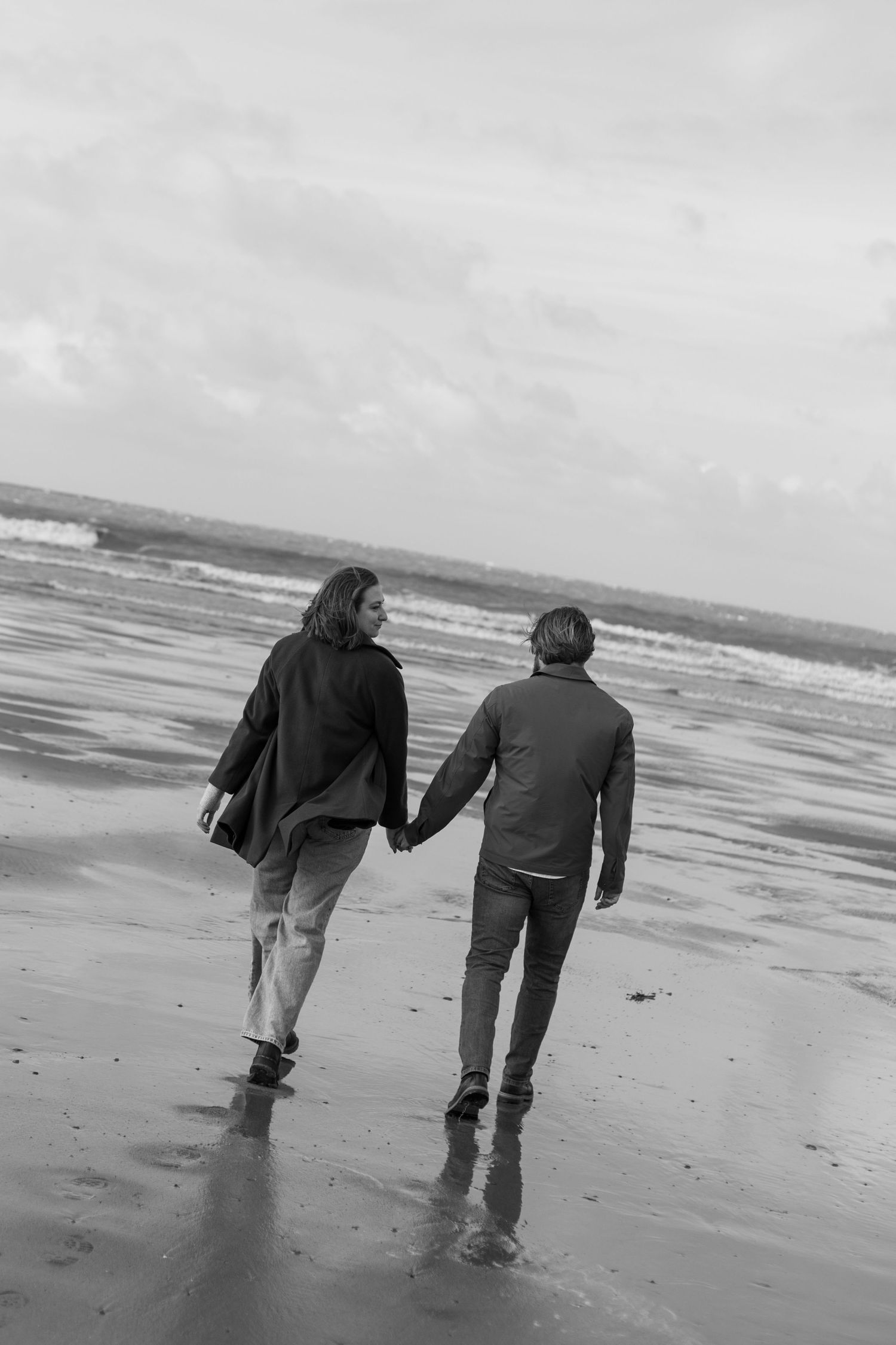 A couple walks hand in hand along a misty beach shore in black and white.