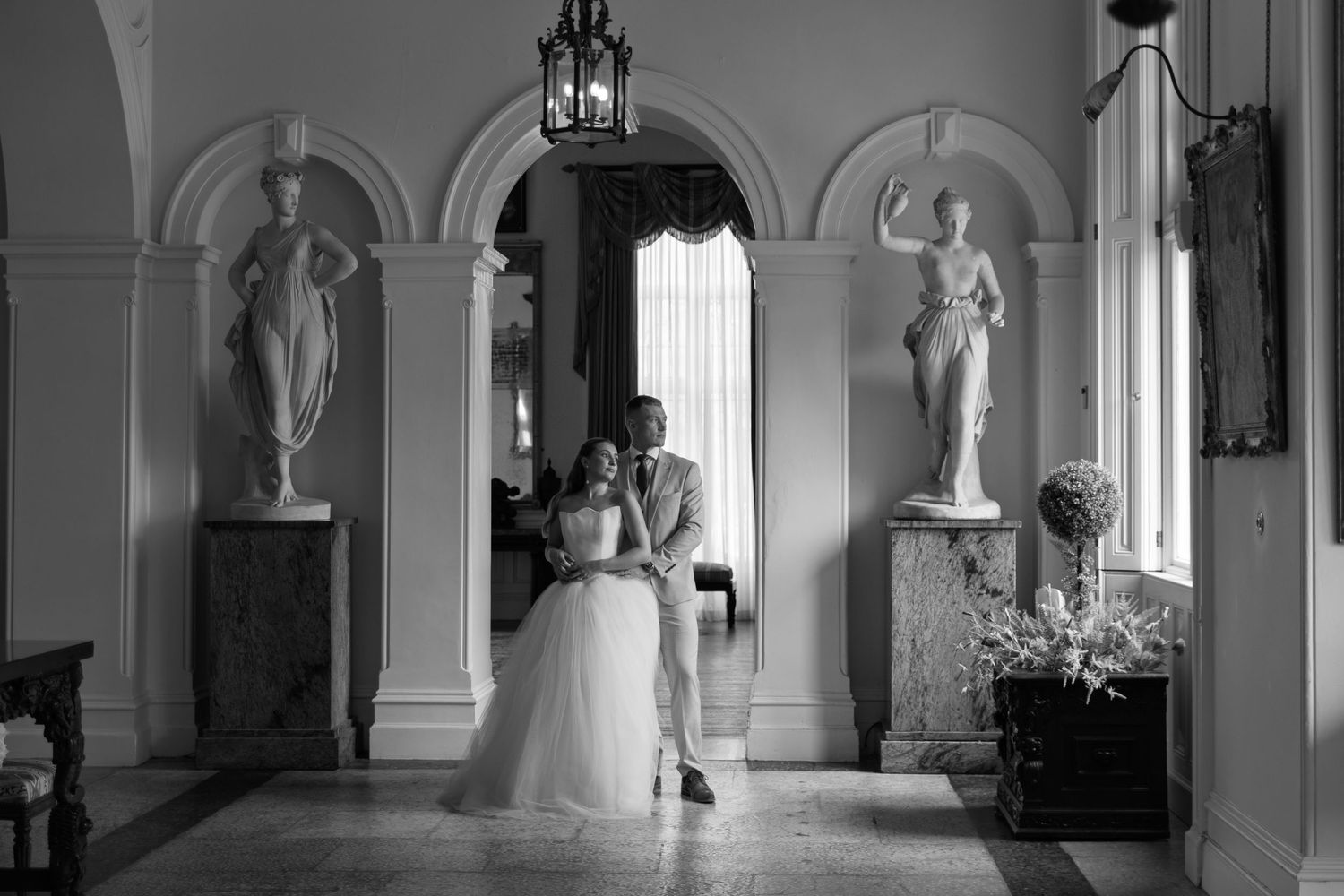 Black and white panoramic view of an elegant corridor with arched windows, columns and classical architectural details with bride and groom.