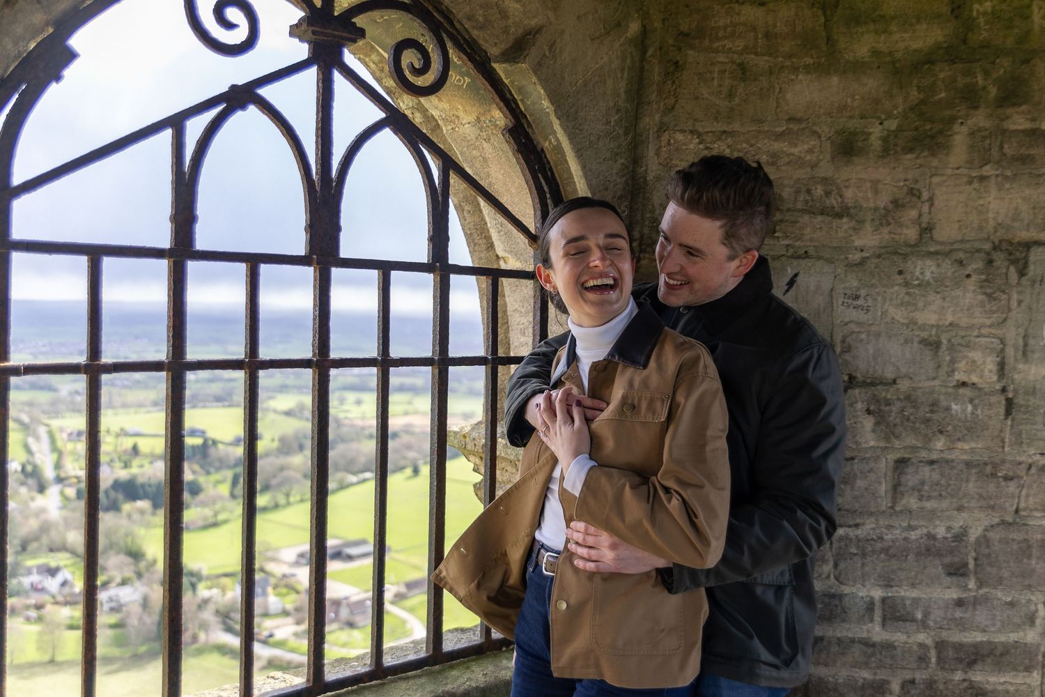 Two people sit on stone steps overlooking a scenic countryside view through arched openings in a historic structure.