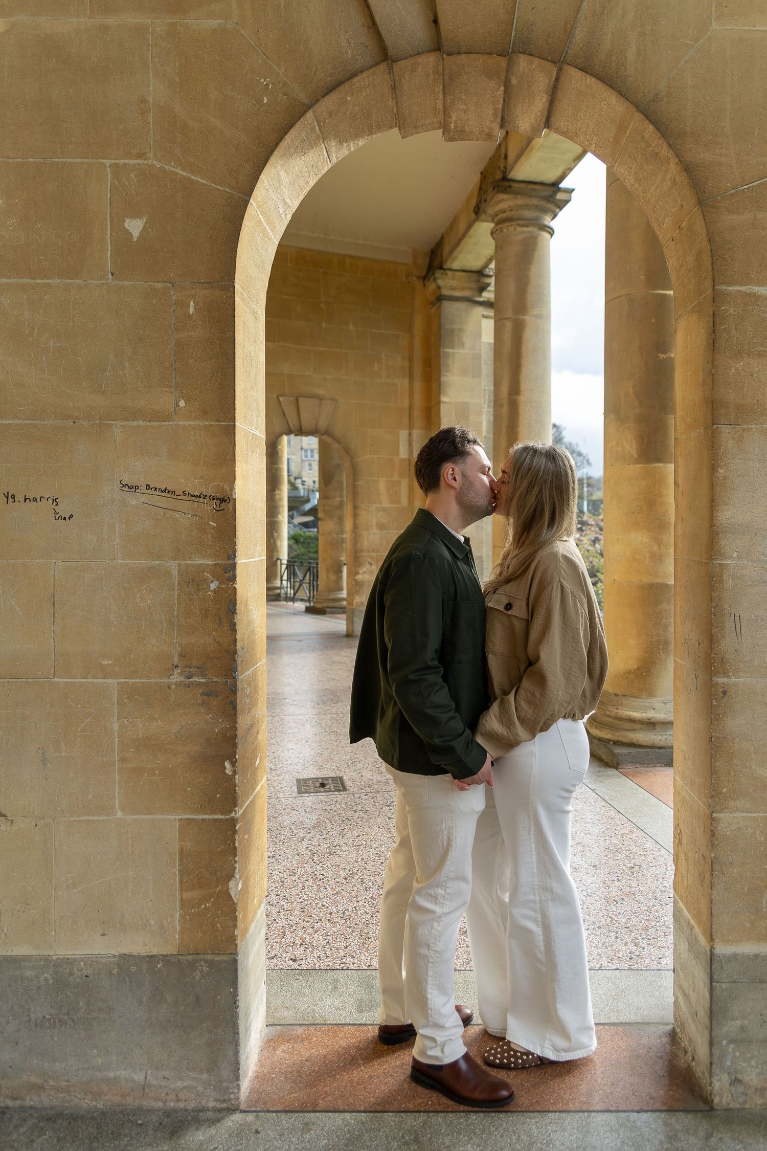 Couple sharing a romantic kiss under a stone archway in a historic building corridor.