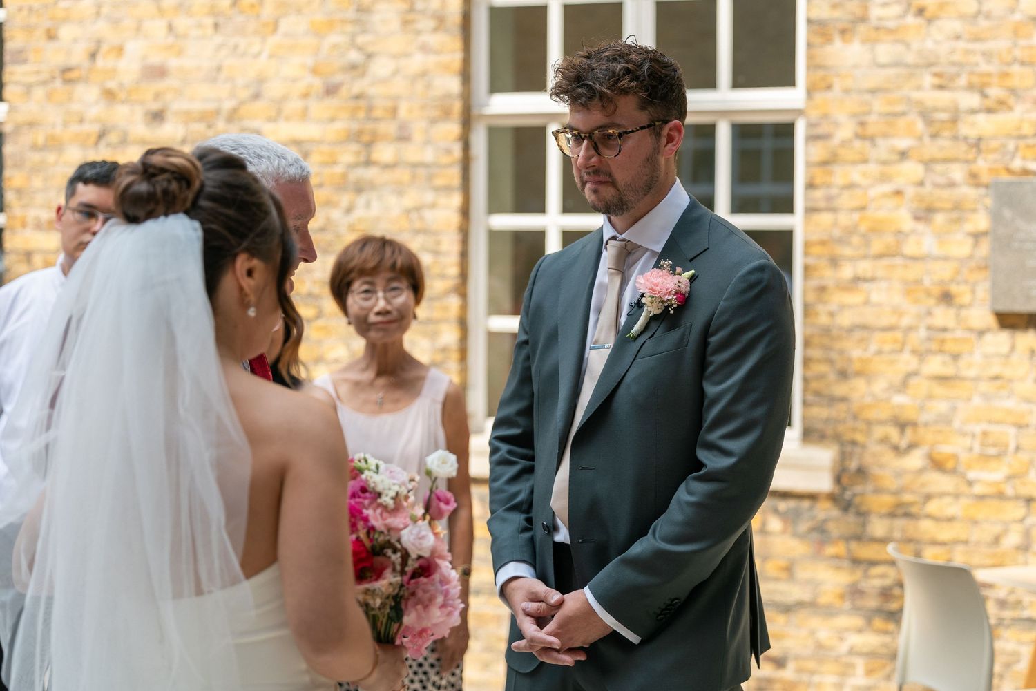 Groom in gray suit with pink boutonniere stands at outdoor wedding ceremony facing bride in white dress and veil.