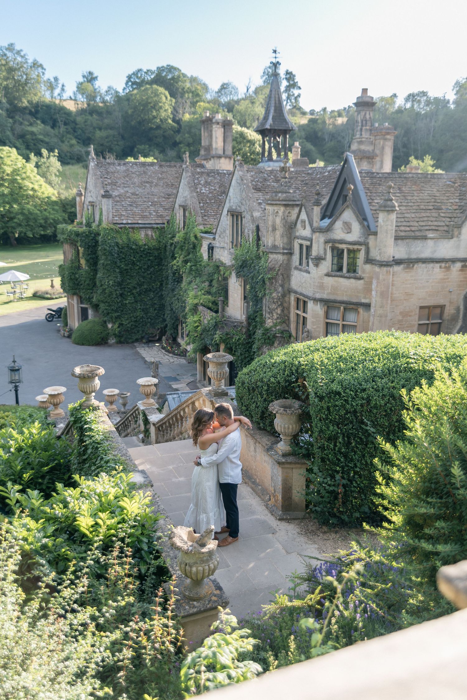 Couple embracing on terrace of ivy-covered English manor house surrounded by lush gardens and stone urns.