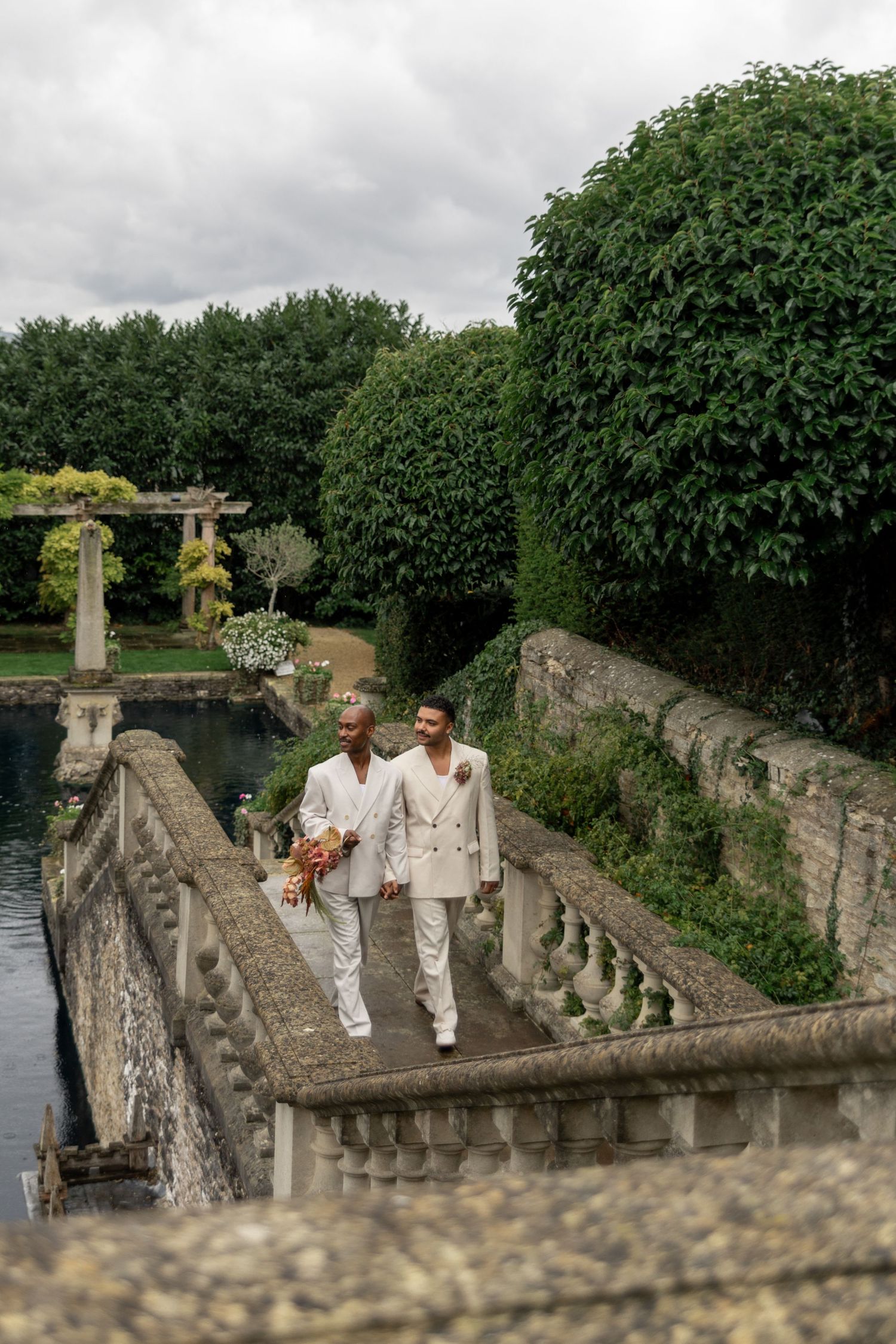 Couple in matching white outfits walk along ornate stone balustrade beside garden pond.