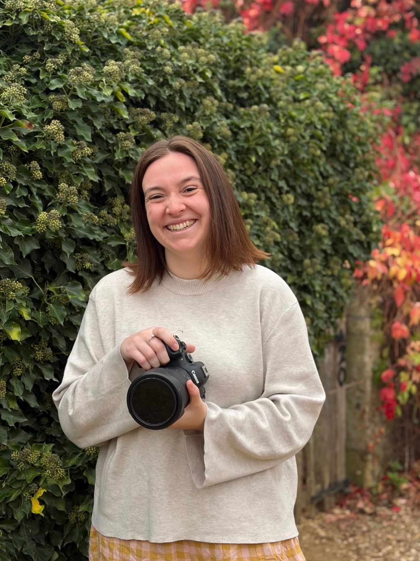 Smiling woman holding a camera outdoors, wearing a beige sweatshirt with colorful autumn foliage in the background.