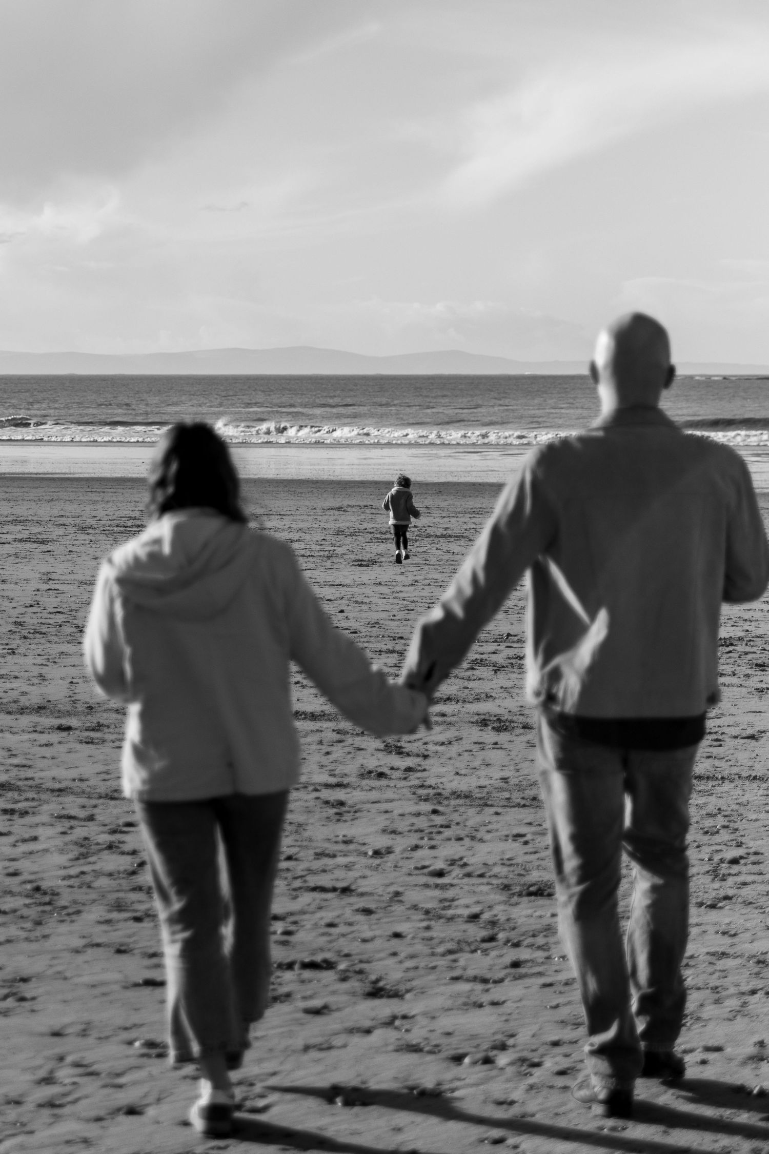 Two figures walk hand in hand along a misty beach shoreline in black and white.