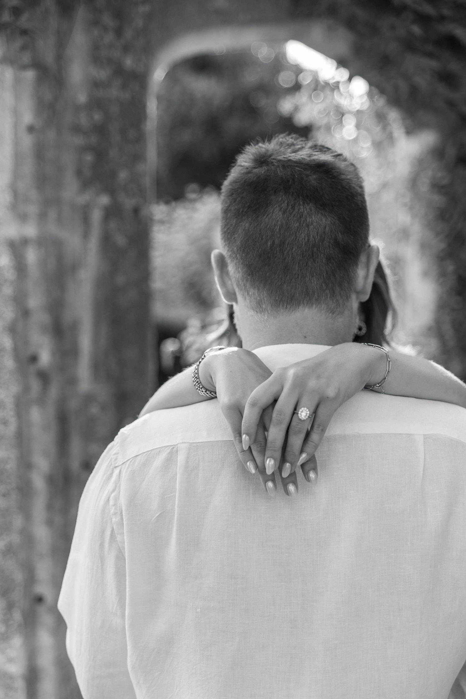 Black and white photo of a couple embracing, woman's hand with engagement ring resting on man's shoulder.