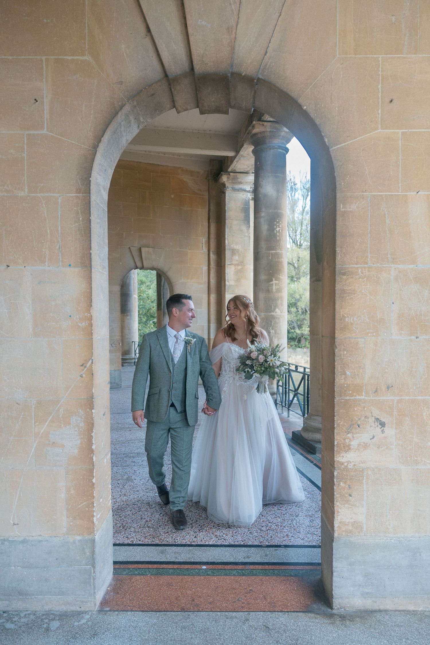 Bride and groom walking hand-in-hand through a stone archway at Parade Gardens, Bath, bride holding a green bouquet.