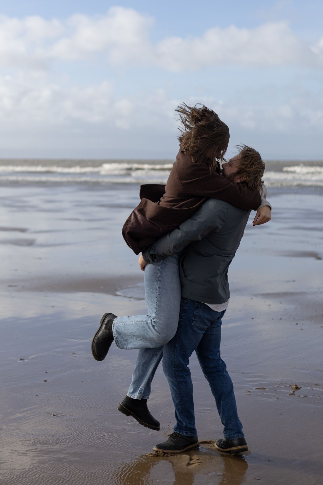 Friends support each other while walking along the shoreline on a cloudy beach day.