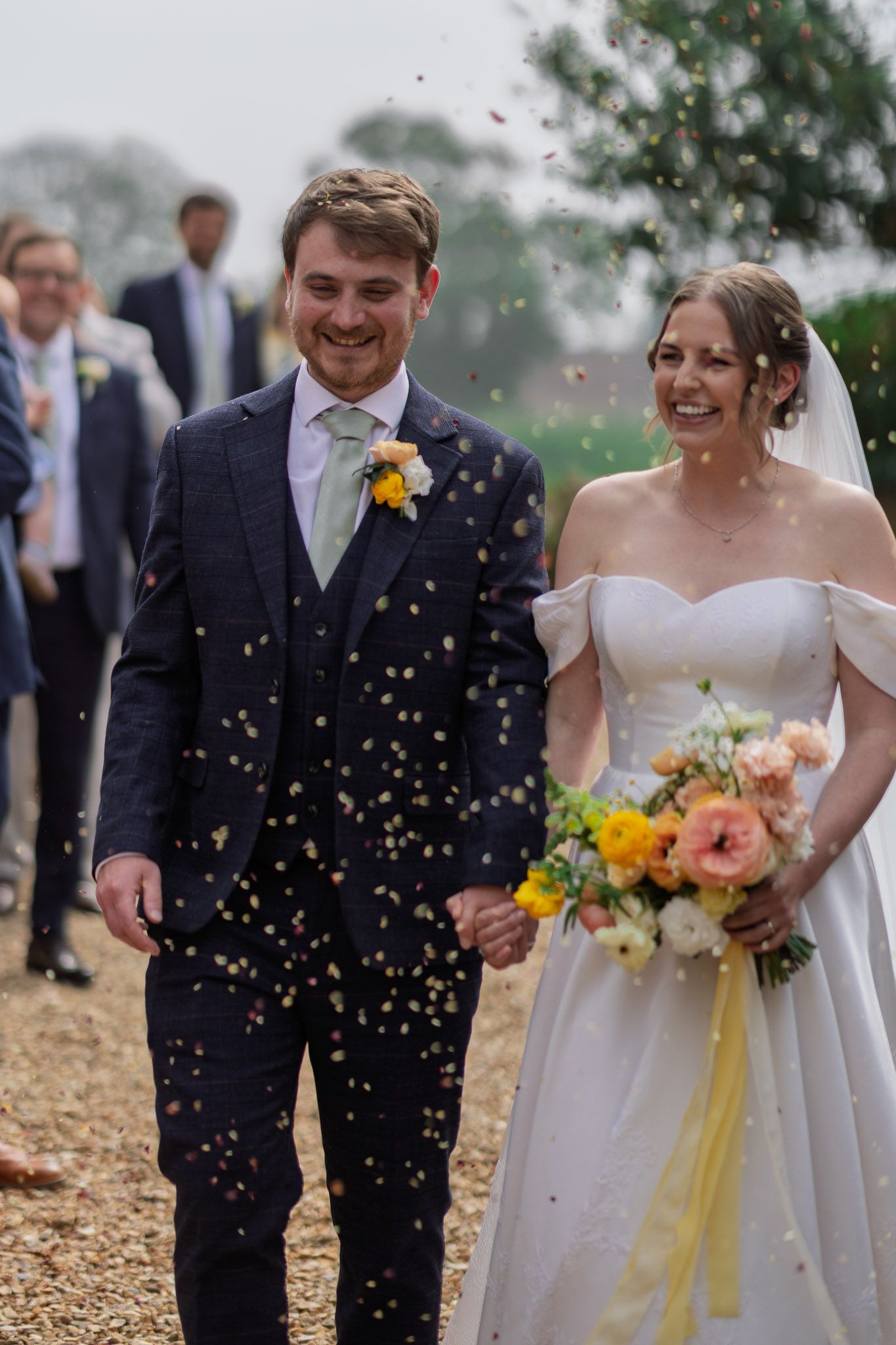 Confetti falls around a happy couple in wedding attire on a gravel pathway.