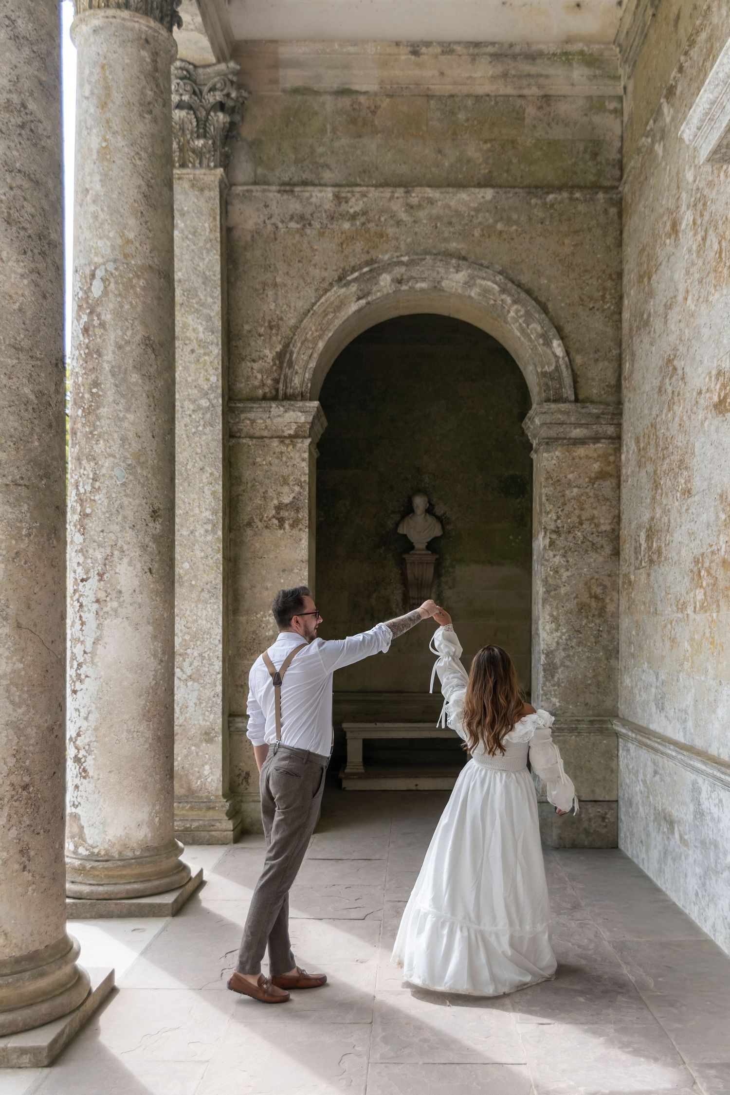 Groom twirls bride in white dress among stone columns in an elegant historic venue with arched alcove.