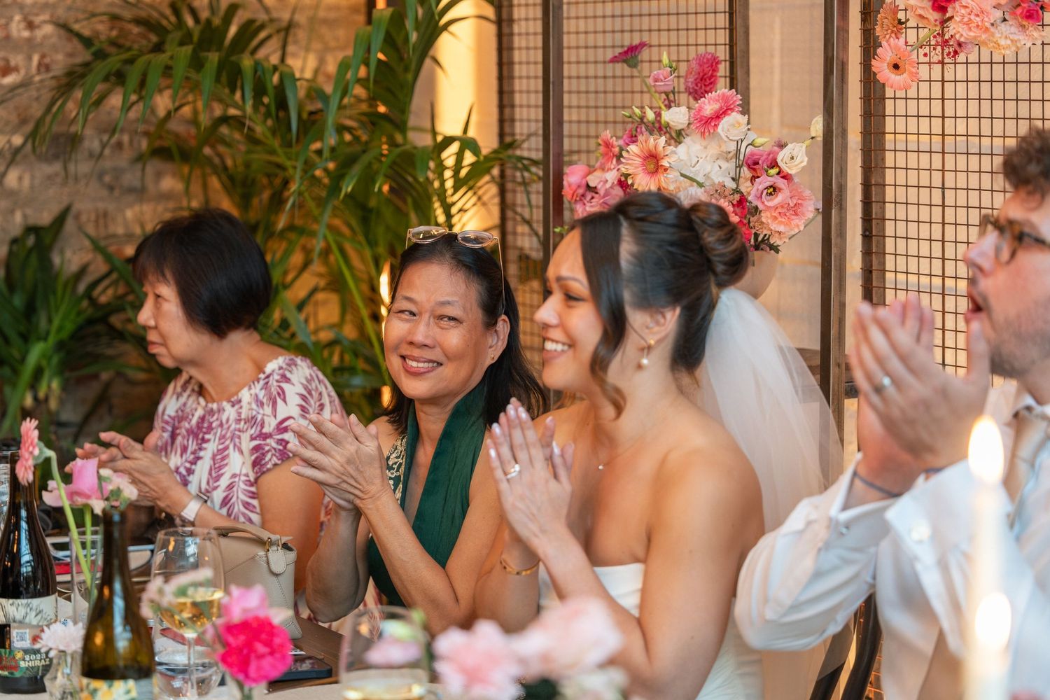 Bride in floral crown applauds with smiling Asian women guests at elegant pink-themed wedding reception dinner.