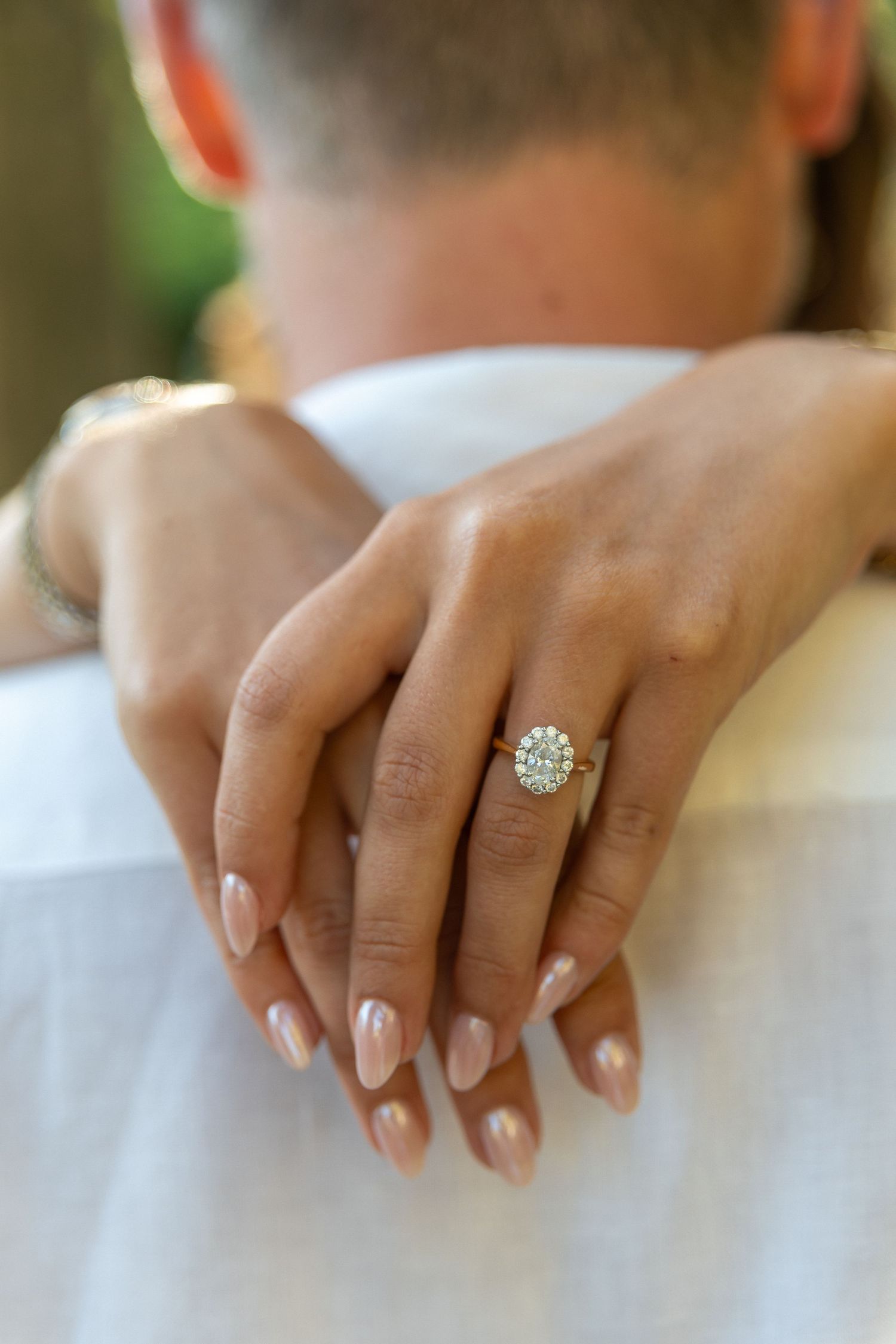 Close-up of a woman's hand with pink nails and diamond halo engagement ring resting on a man's shoulder.