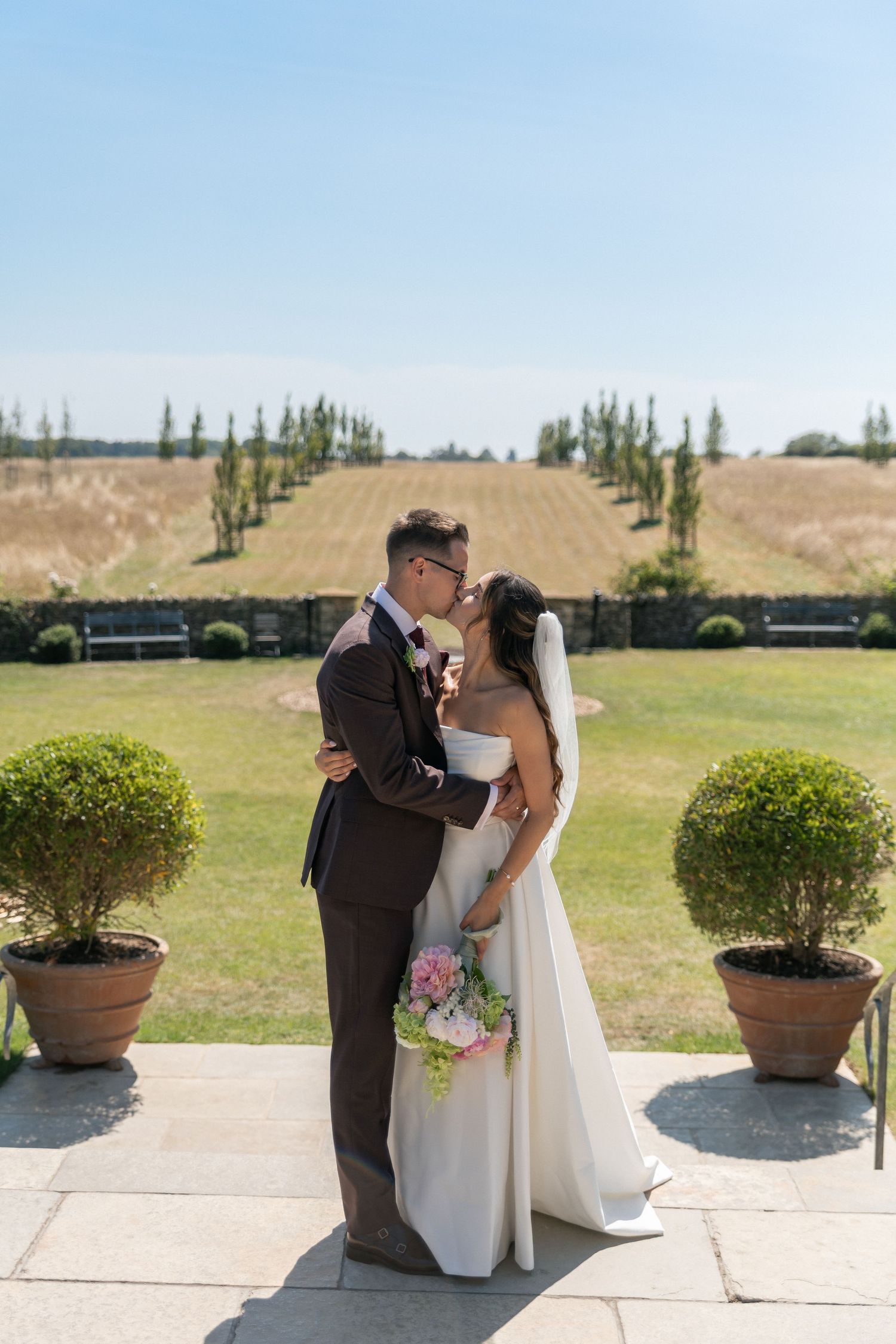 Bride and groom share a romantic kiss on a terrace, with manicured gardens and tall trees in the background on a sunny day.
