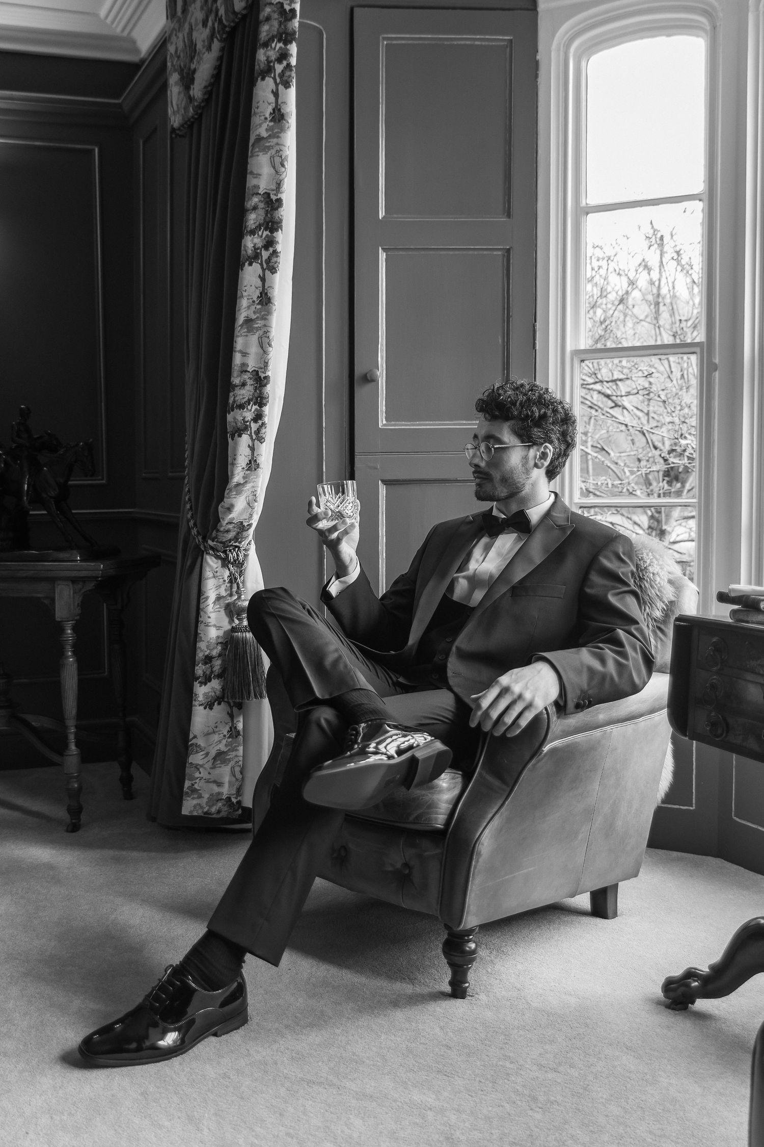 Black and white photo of an elegant man in a suit sitting in an armchair holding a whiskey glass near a window.