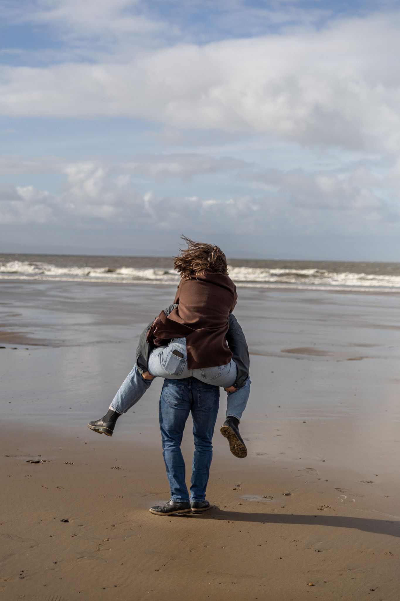 Person in jeans and dark jacket bending down playfully on a windy beach with waves in the background.